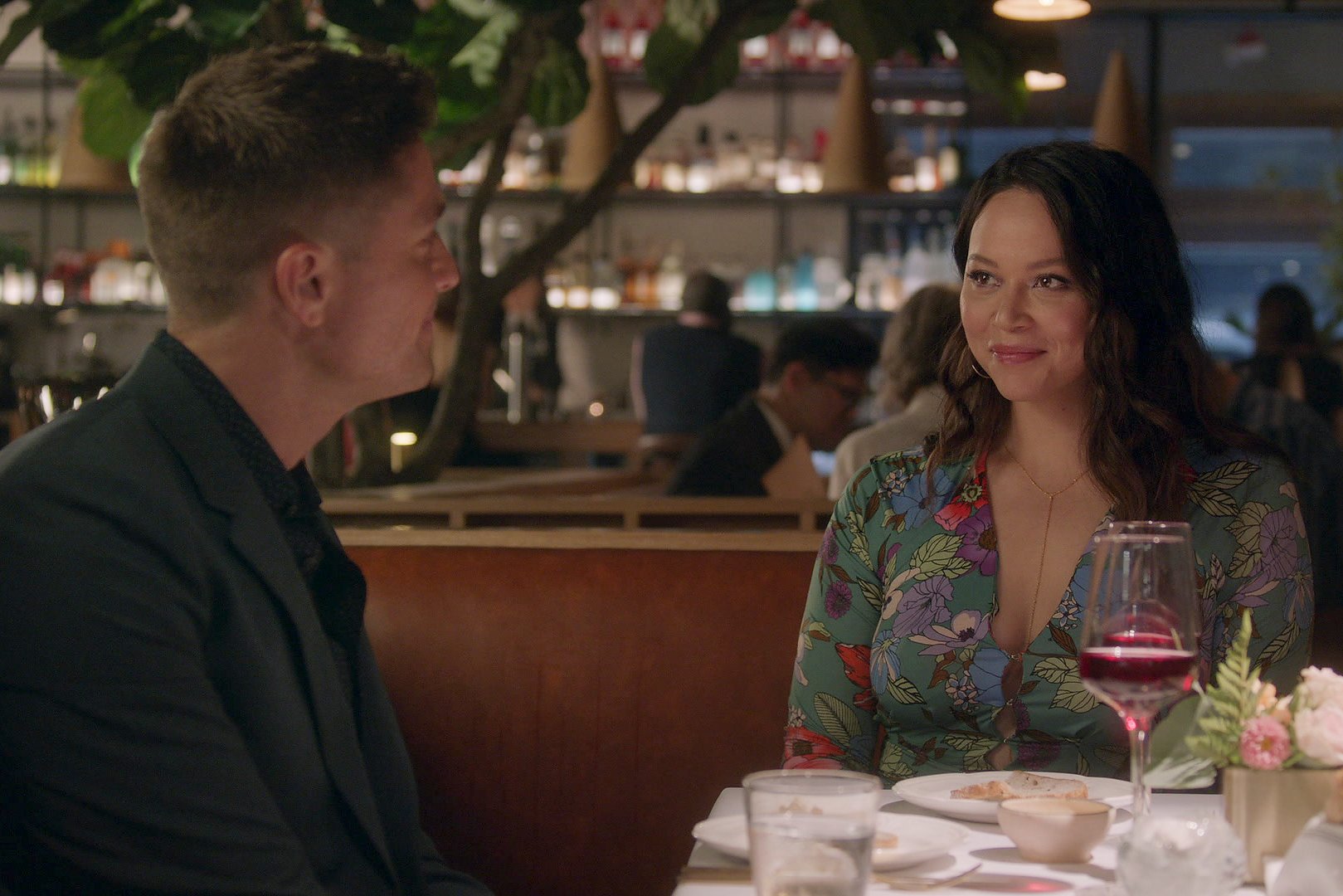 Eric Winter and Melissa O'Neil, in character as Tim Bradford and Lucy Chen, share a scene at a restaurant in 'The Rookie' Season 5 Episode 10, 'The List,' on ABC. Tim wears a black suit. Lucy wears a green dress with multi-colored flowers on it.