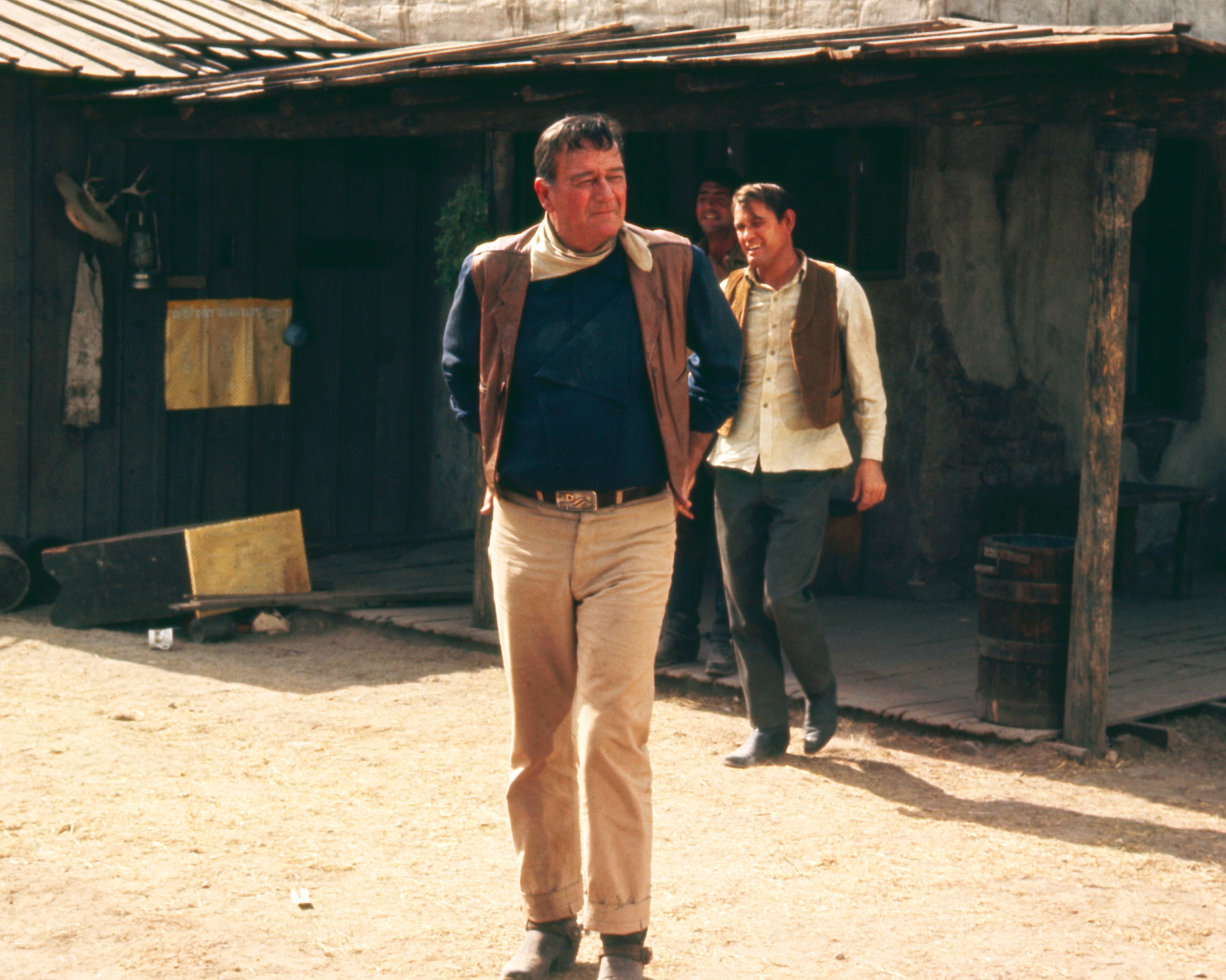 'The Sons of Katie Elder' John Wayne as John Elder and Earl Holliman as Matt Elder in Western pants. They're walking outside of a house on a dirt road.