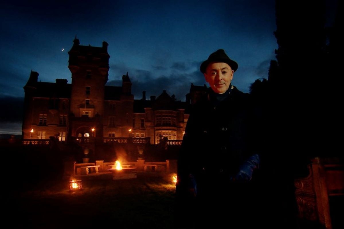 Alan Cumming, the host of 'The Traitors' on Peacock, wears all blackstanding in front of a castle.