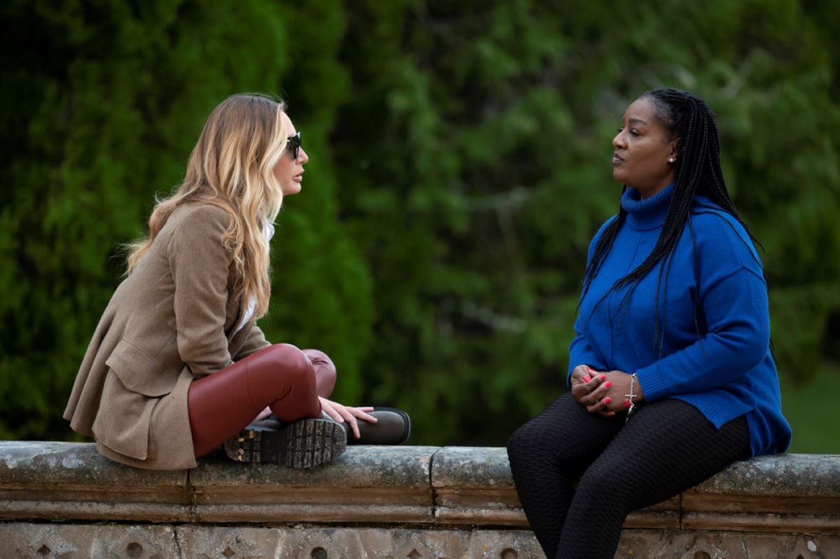 Kate Chastain and Cirie Fields, who, according to 'The Traitors' spoilers, won, sit outside the castle. Kate wears a tan coat, red leggings, and dark brown boots. Cirie wears a royal blue turtleneck sweater and black leggings.