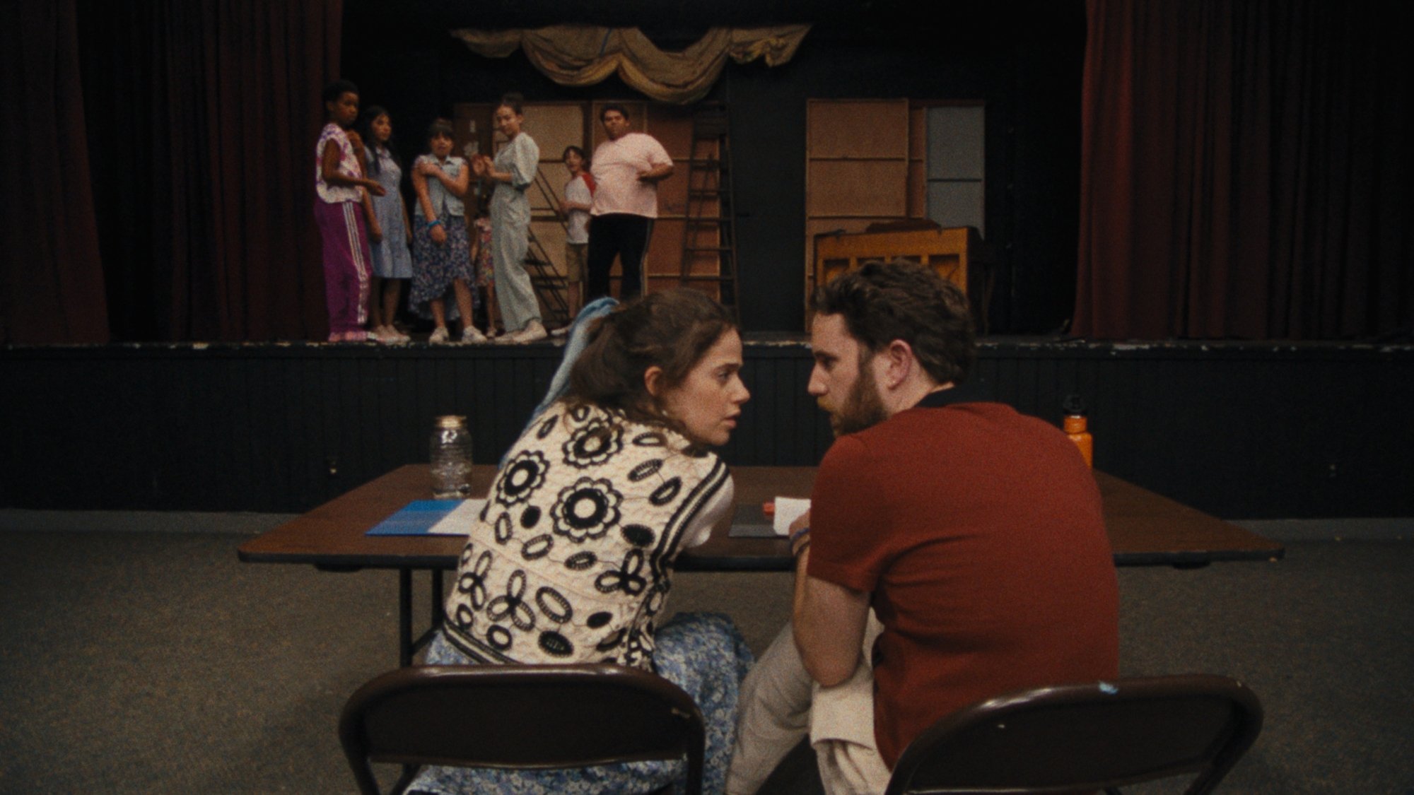 'Theater Camp' Molly Gordon as Rebecca-Diane and Ben Platt as Amos whispering to each other, sitting at a table in front of a stage full of kids.