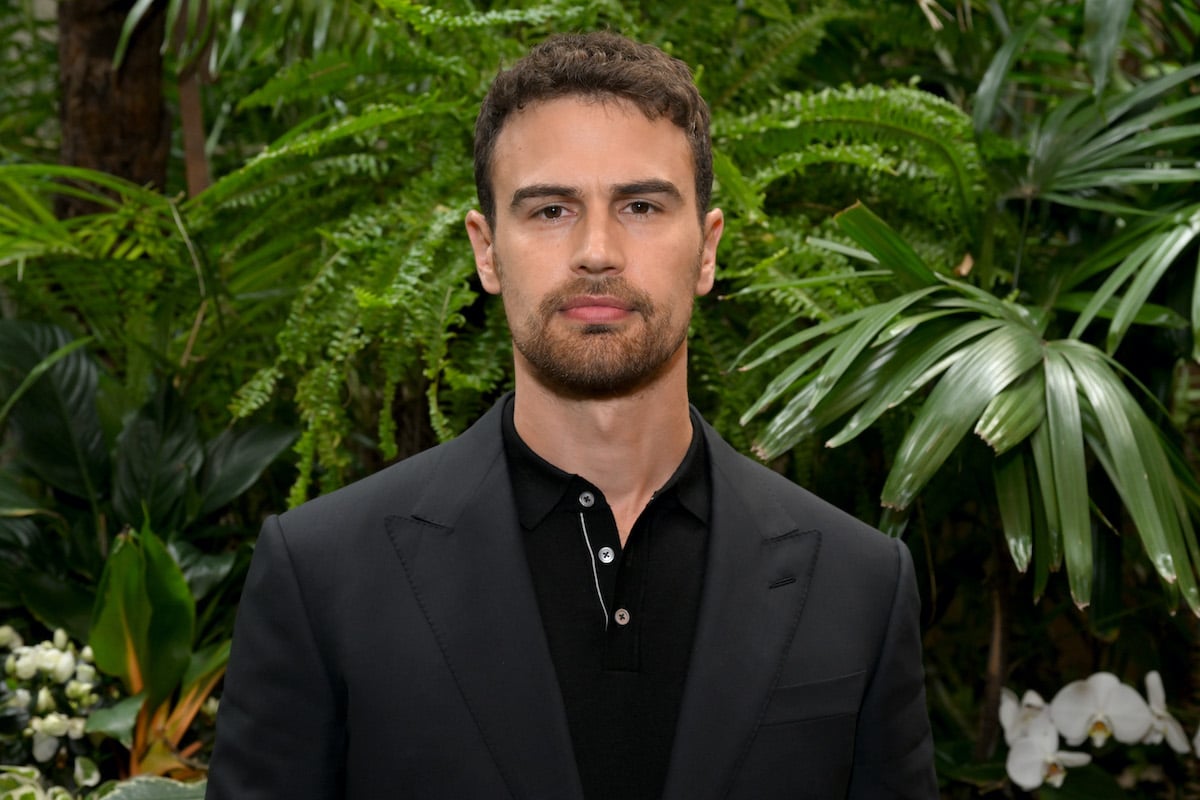 Theo James stands in front of greenery wearing a black suit