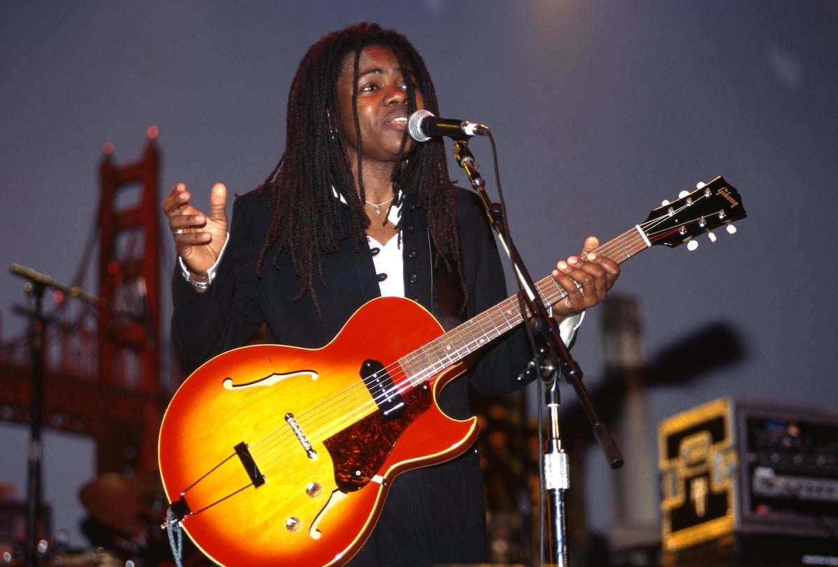 Tracy Chapman sings into a microphone while playing the guitar.