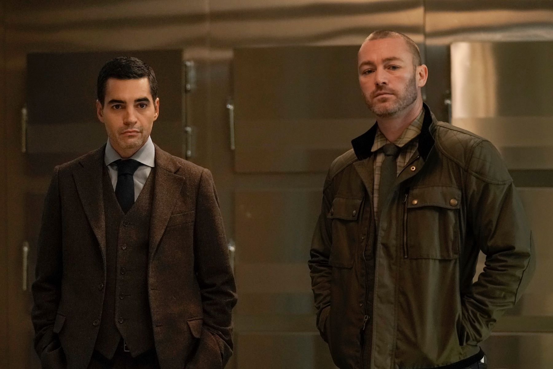 Ramón Rodríguez and Jake McLaughlin, in character as Will Trent and Michael Ormewood, share a scene in 'Will Trent' Season 1 Episode 4. Will wears a dark brown three piece suit over a light gray button-up shirt and black tie. Ormewood wears a dark green jacket over a light green plaid shirt and dark green tie.