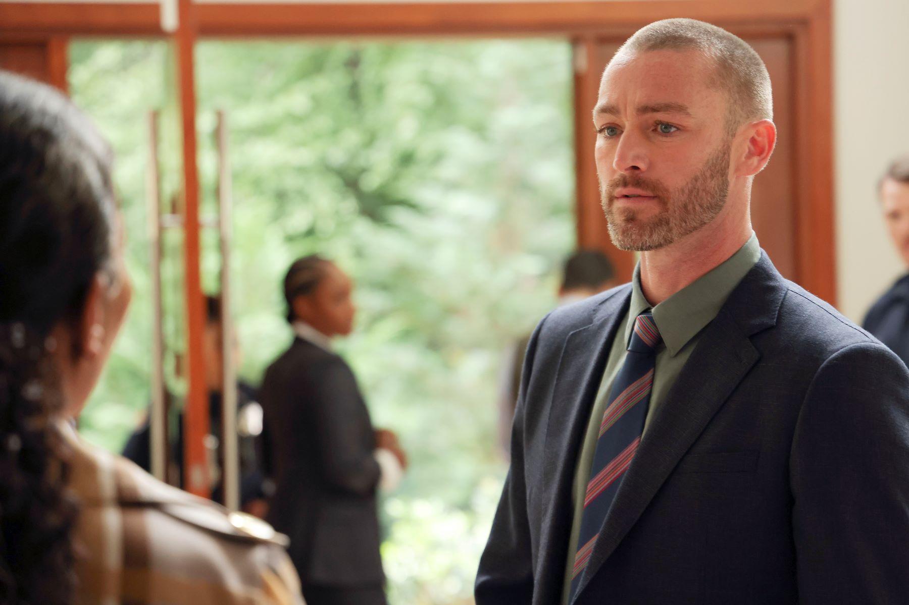 Jake McLaughlin, in character as Michael Ormewood in 'Will Trent' Season 1 Episode 1, wears a dark blue suit over a green button-up shirt and blue, red, and tan striped tie.