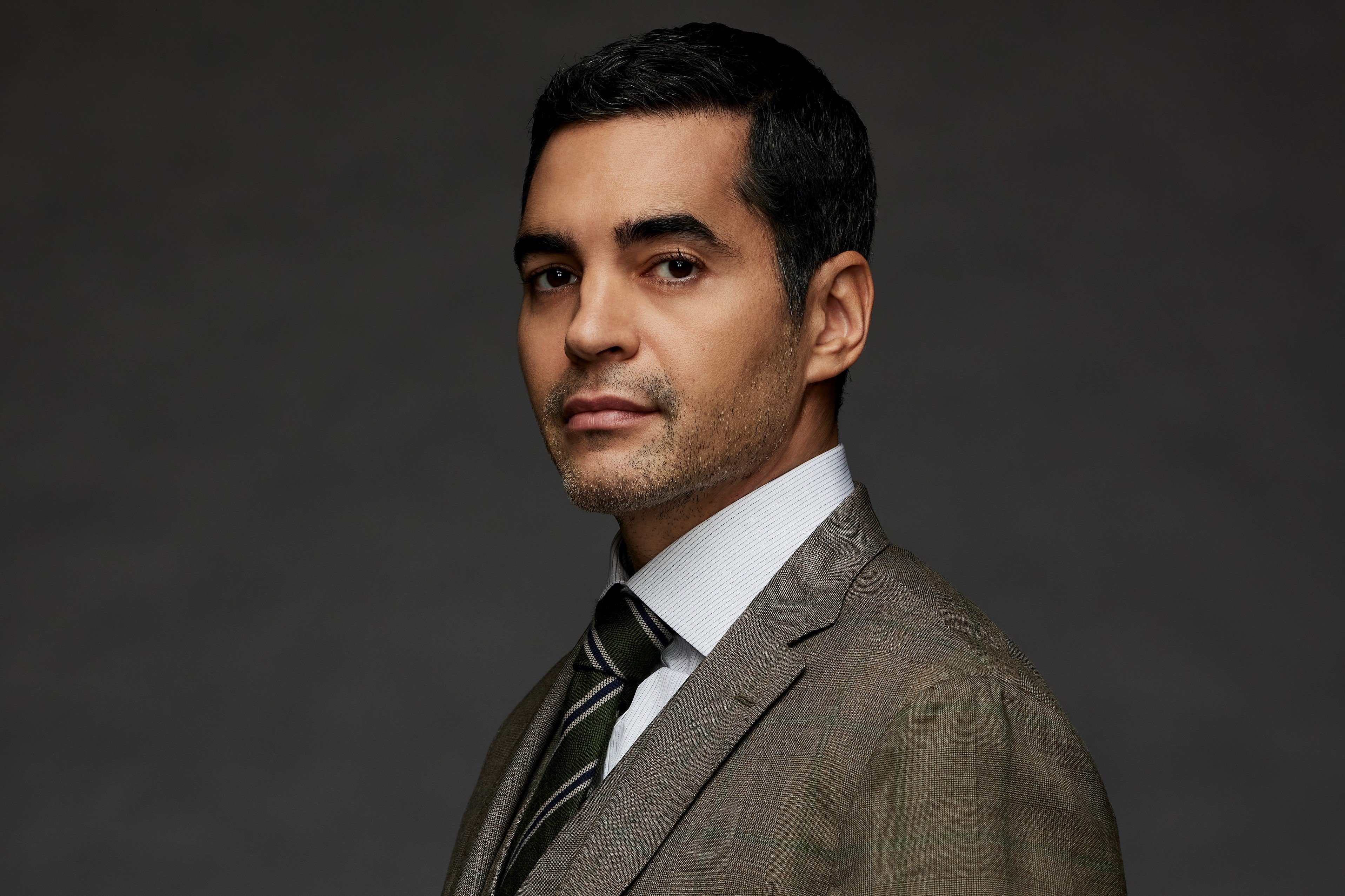Ramón Rodríguez, in character as Will Trent, wears a tan suit over a white button-up shirt and dark green, gray, and black tie.