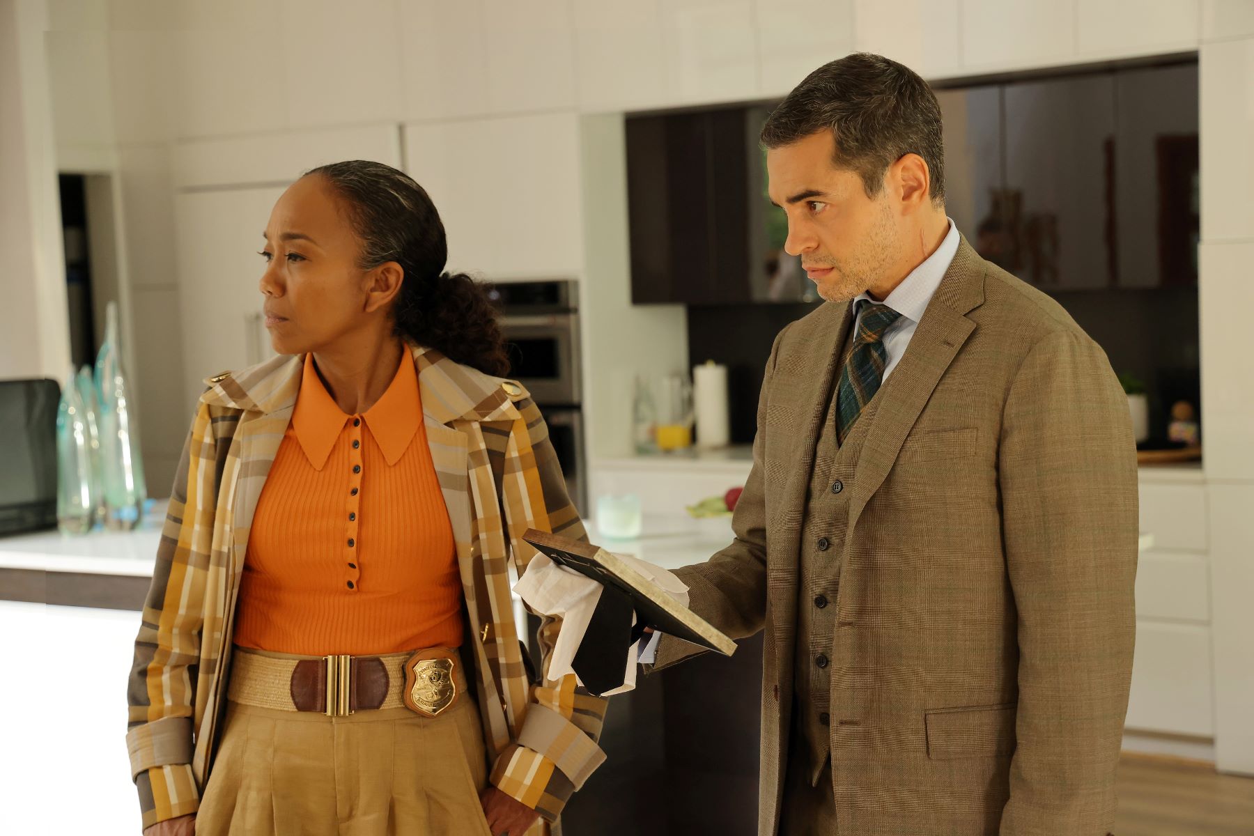 Sonja Sohn and Ramón Rodríguez, in character as Amanda Wagner and Will Trent, in the 'Will Trent' series premiere. Amanda wears a brown and light orange plaid coat over an orange shirt and tan pants. Will wears a brown three-piece suit with a dark green plaid tie.