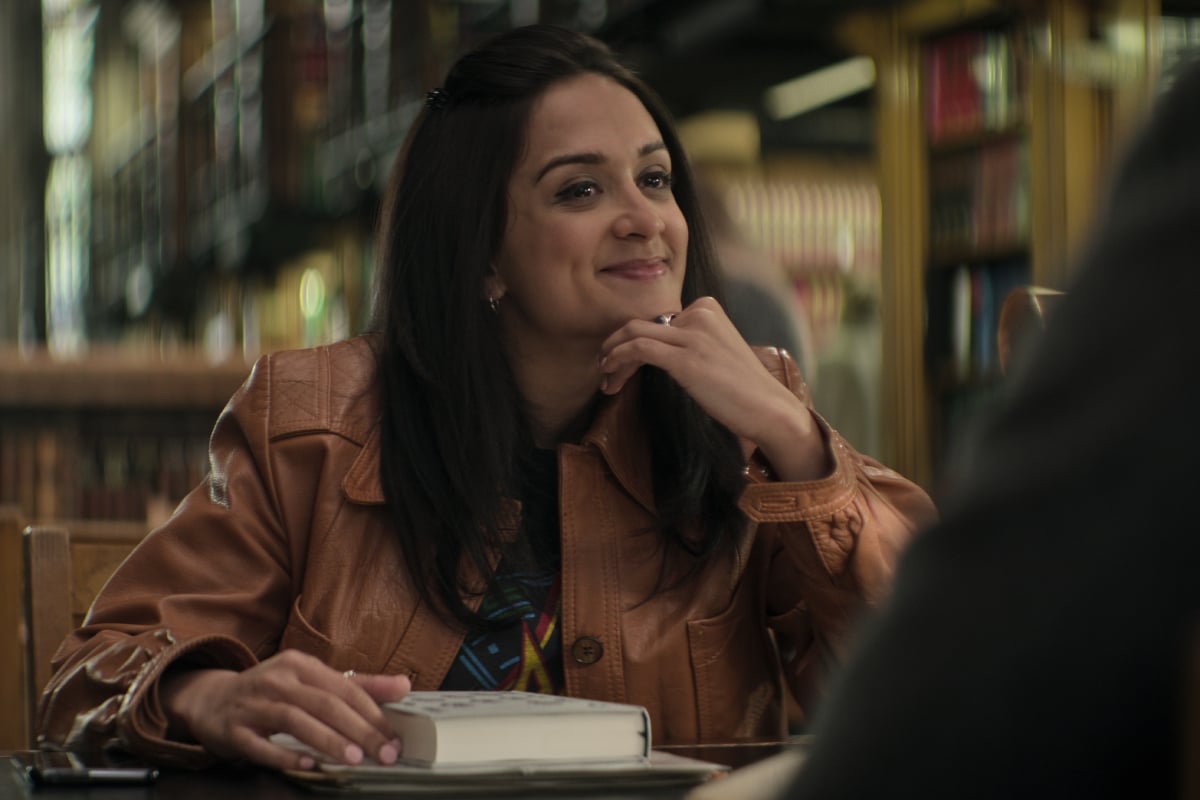 Amy-Leigh Hickman as Nadia Farran in You Season 4. Nadia sits at a table at the library wearing a tan leather jacket.
