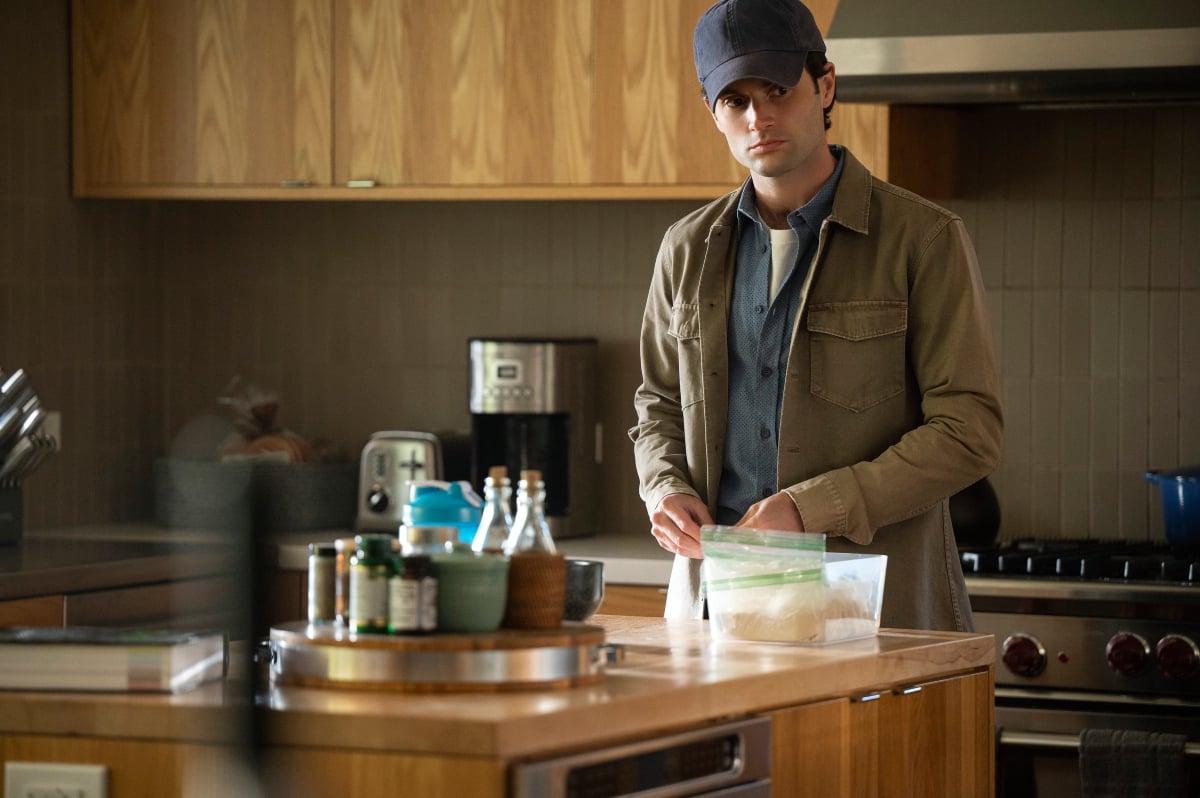 In You Season 4, Joe brings along his favorite disguise. Joe stands in a kitchen wearing a baseball cap, button-down shirt, and tan jacket.