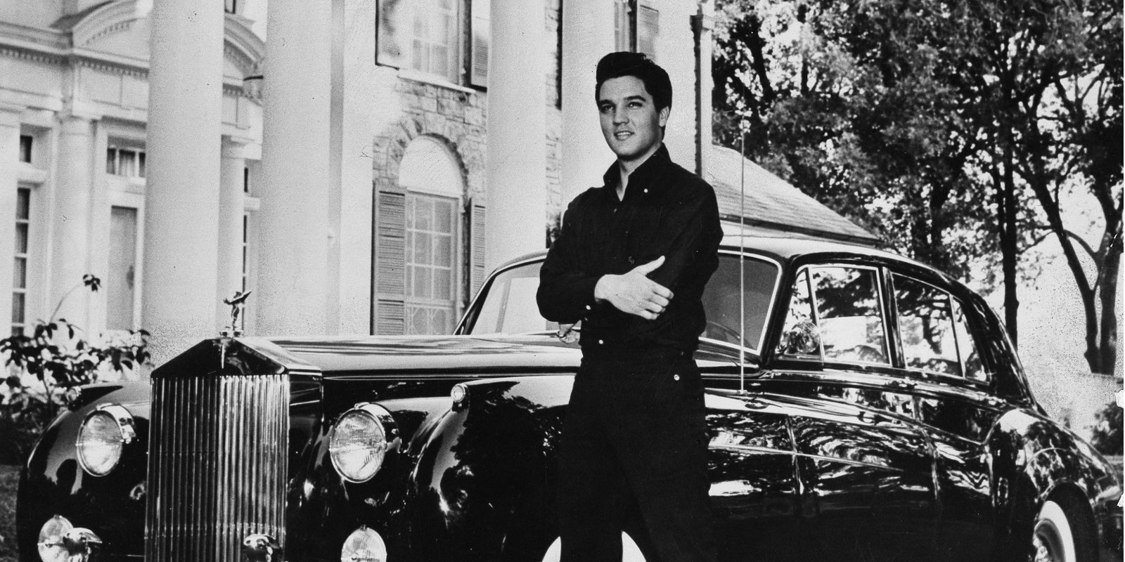 Elvis Presley poses in front of Graceland in a black and white image.