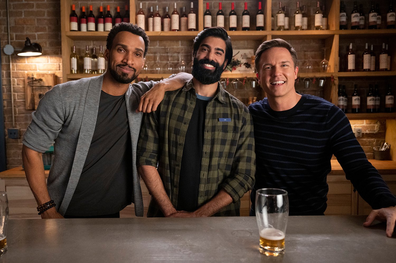 Ginny & Georgia stars Nathan Mitchell (Zion), Raymond Ablack (Joe), and Scott Porter (Paul) pose together behind the bar at Blue Farm.