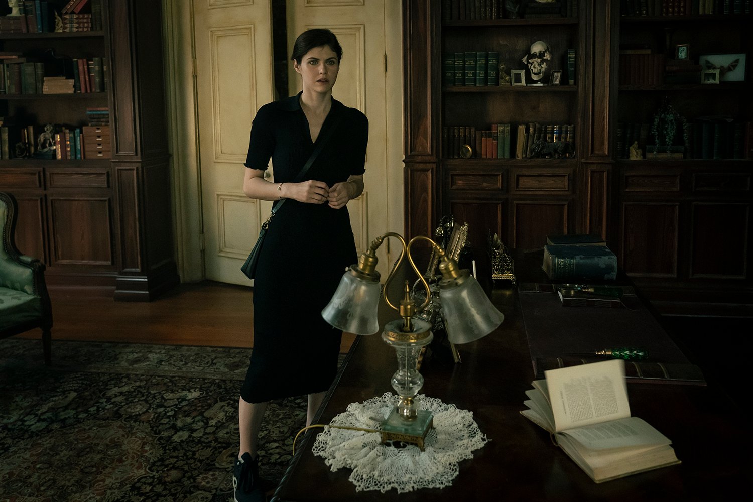 Mayfair Witches star Alexandra Daddario as Rowan Fielding standing by a desk in a library while wearing a black dress.