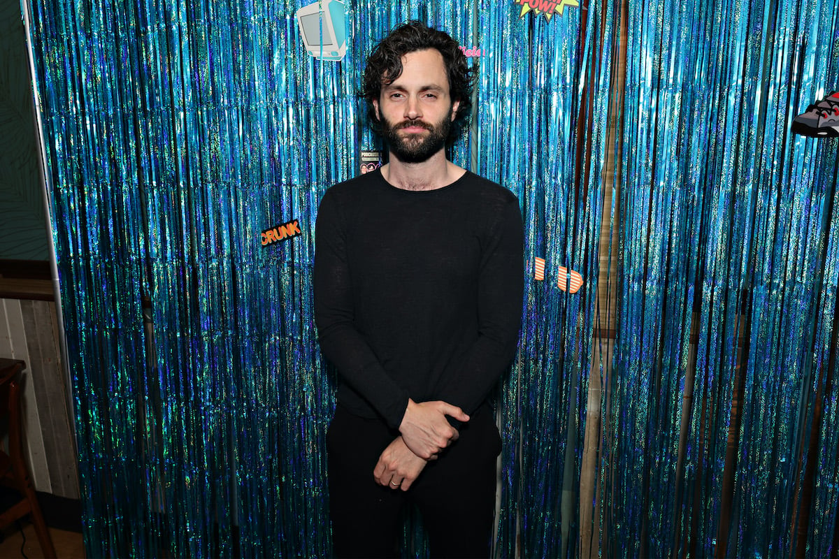 name actor Penn Badgley stands in front of blue streamers in a black shirt