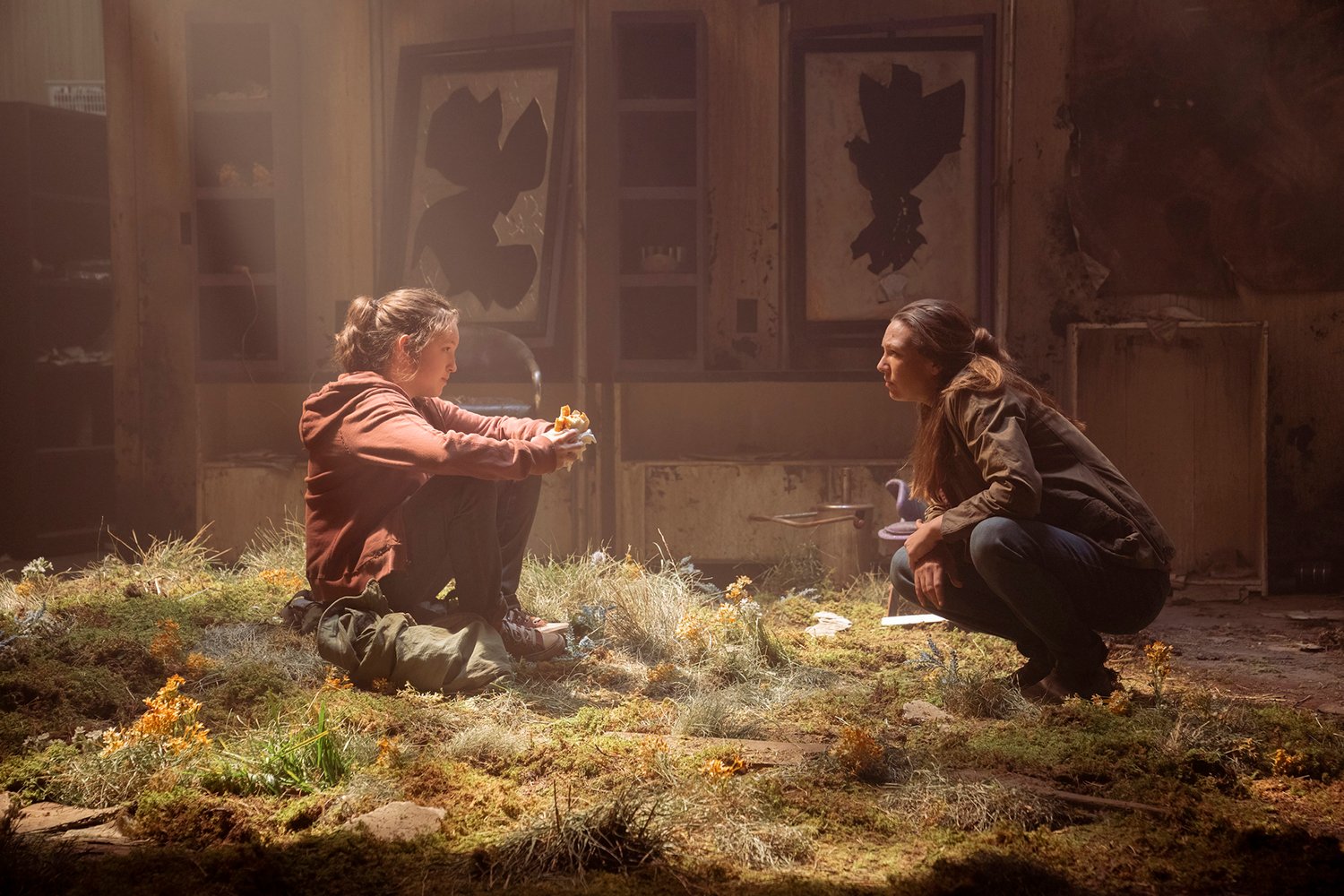 The Last of Us: Bella Ramsey as Ellie sitting on the grass under a beam of light as Anna Torv's Tess kneels in front of her
