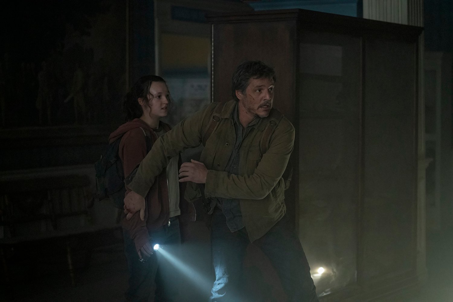 Watch The Last of Us: Bella Ramsey as Ellie holding a flashlight down while Pedro Pascal as Joel blocks her with his arms.