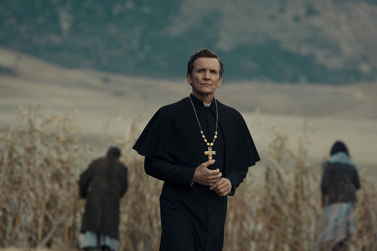 In 1923 Episode 8, Father Renaud stands in a field wearing a black cassock and cross around his neck.