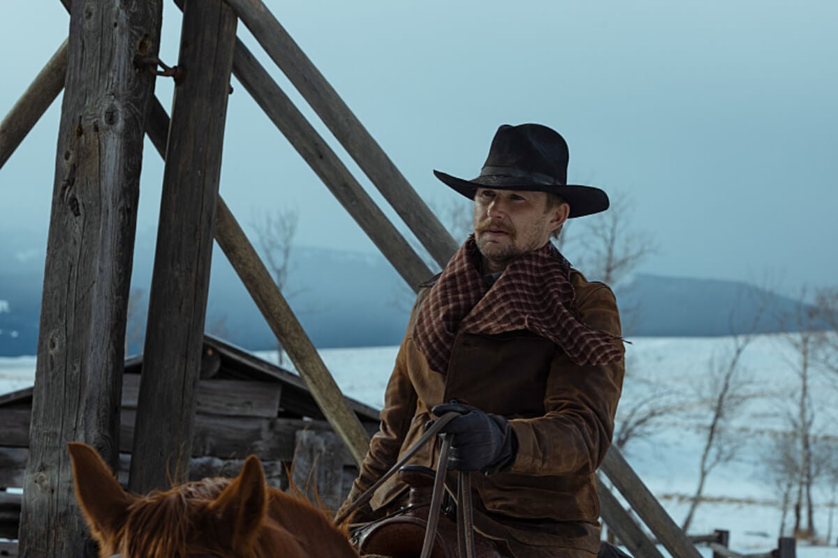 In 1923, Zane rides a horse through the snow. He is wearing a brown coat, a scarf, and a black hat.