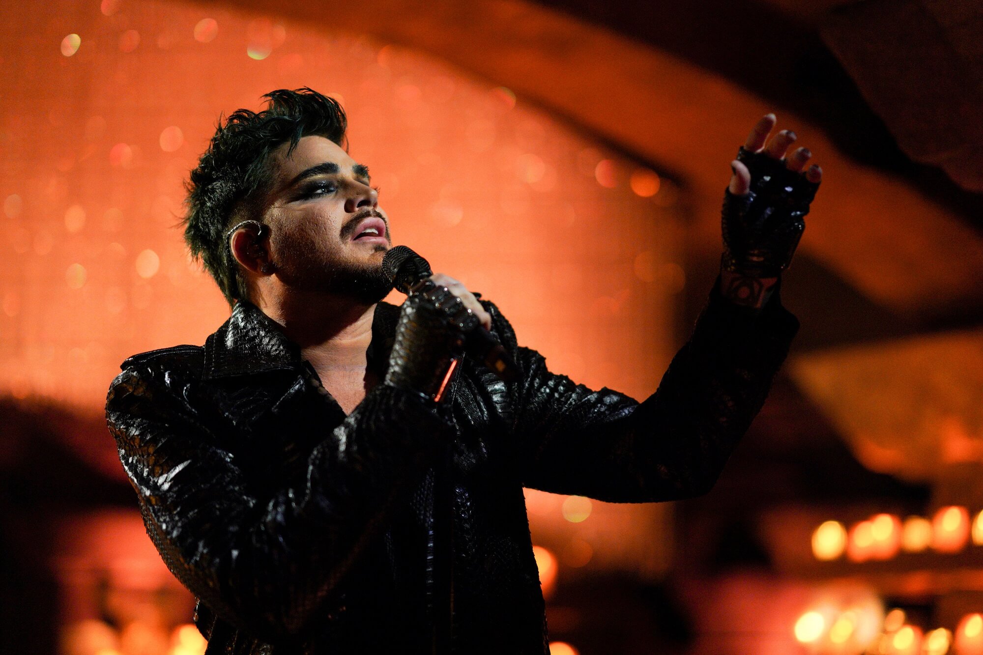 Adam Lambert performs wearing a black outfit with orange lighting behind him