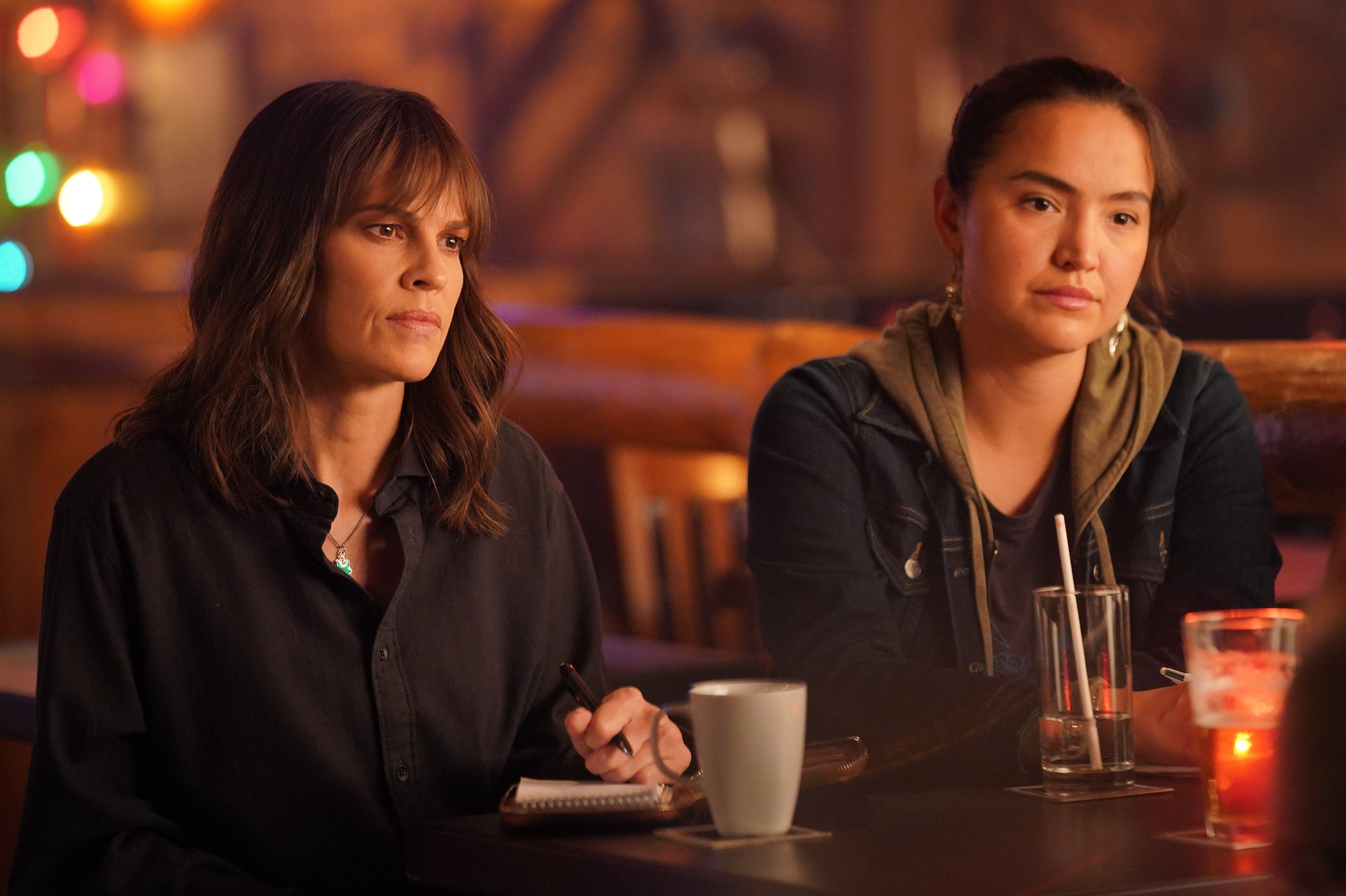 Hilary Swank and Grace Dove, who star as Eileen Fitzgerald and Roz Friendly in episodes of 'Alaska Daily' Season 1, share a scene at a bar. Eileen wears a black button-up shirt. Roz wears a dark jean jacket over an olive green hoodie and dark gray shirt.