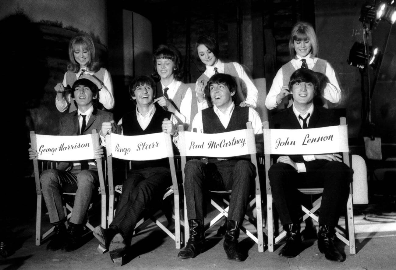 A black and white picture of The Beatles sitting in director's chairs in front of co-stars on 'A Hard Day's Night.'