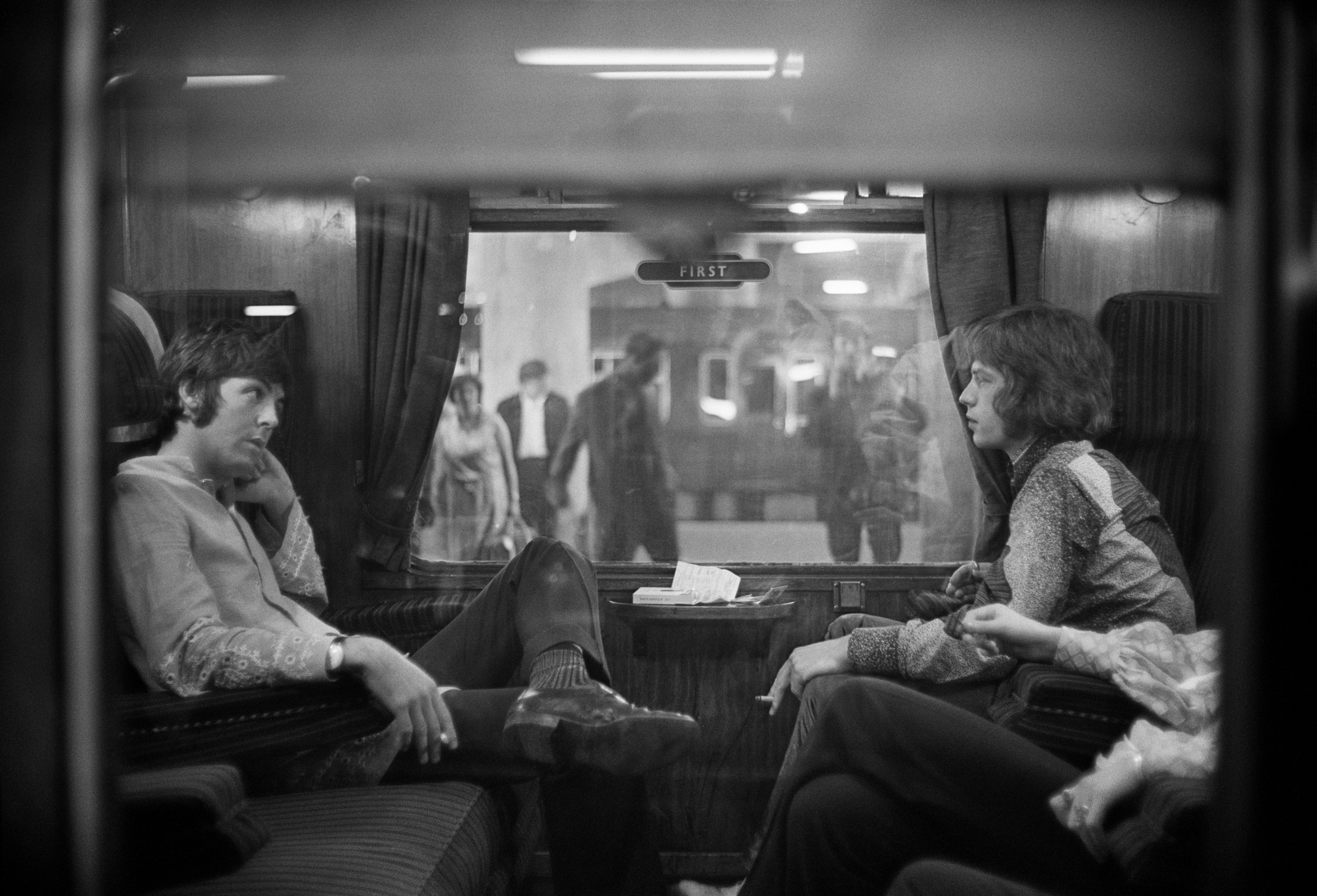 Paul McCartney of The Beatles and Mick Jagger of The Rolling Stones sit across from each other on a train