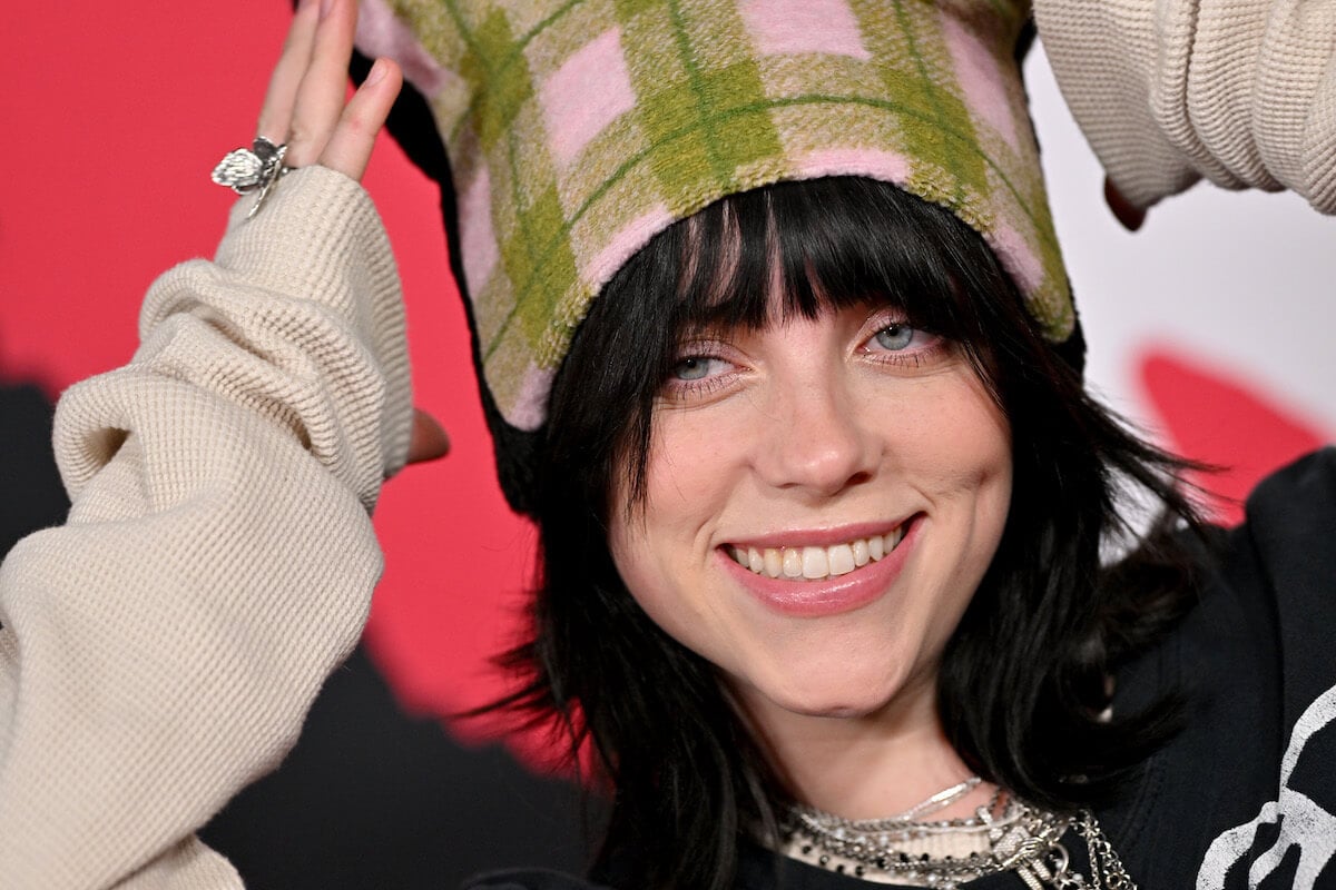 Singer Billie Eilish smiles wearing a plaid beanie and chain necklaces.