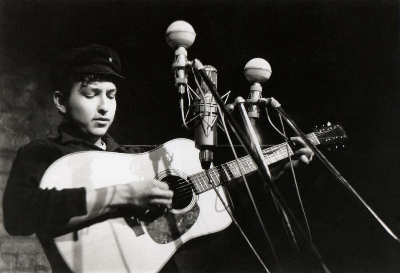 A black and white picture of Bob Dylan playing guitar in front of three microphones.
