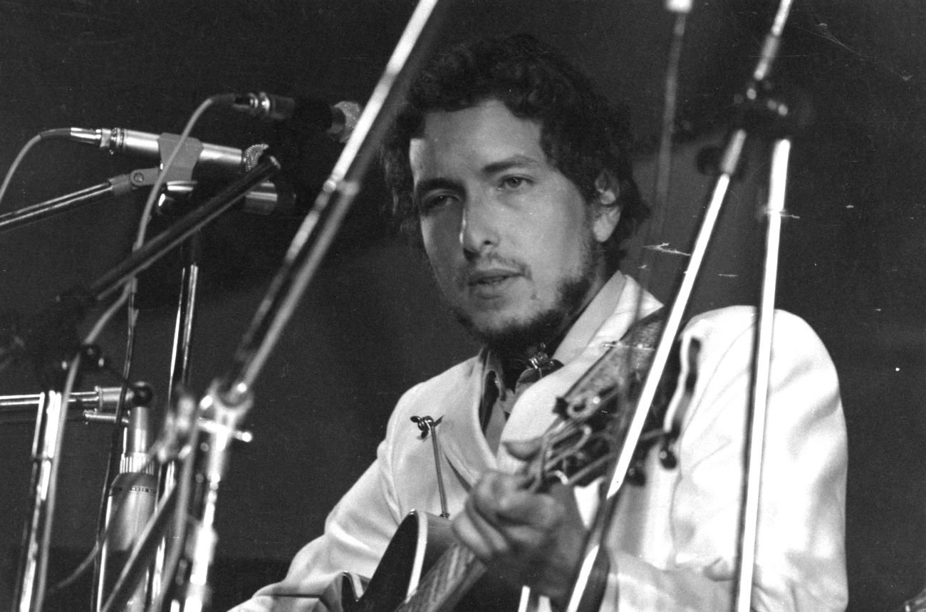 A black and white picture of Bob Dylan playing guitar behind multiple microphones.