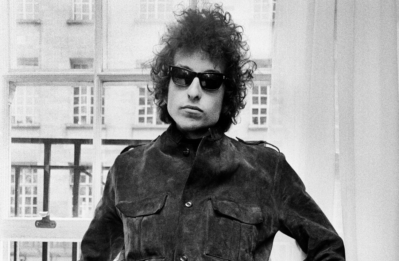 A black and white picture of Bob Dylan wearing glasses and leaning against a window.