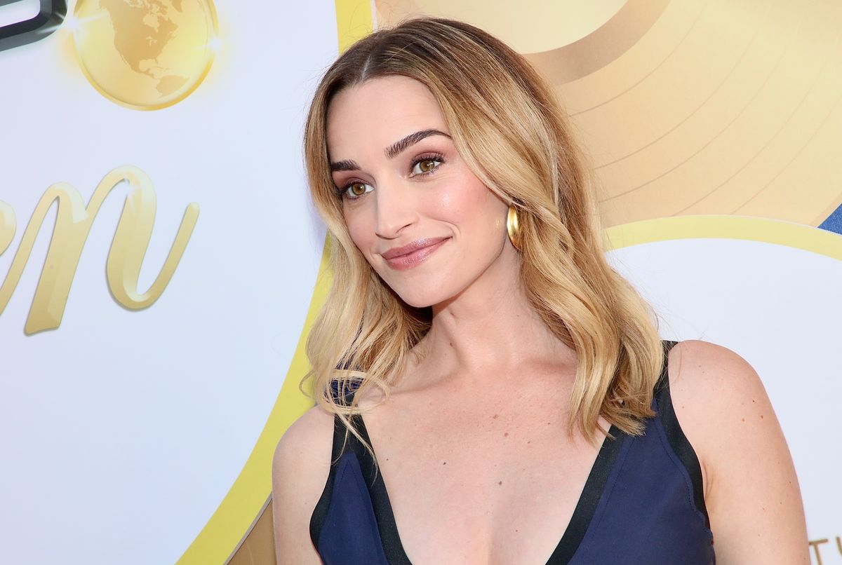 Brianne Howey poses in front of a white and gold backdrop.