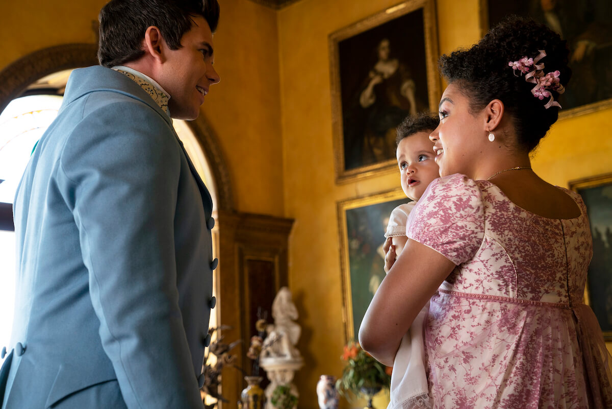 Luke Newton as Colin Bridgerton, Otis, Rocco, and Prince Shillingford as Baby Oliver Crane and Ruby Barker as Marina Thompson chatting in the drawing room in 'Bridgerton'