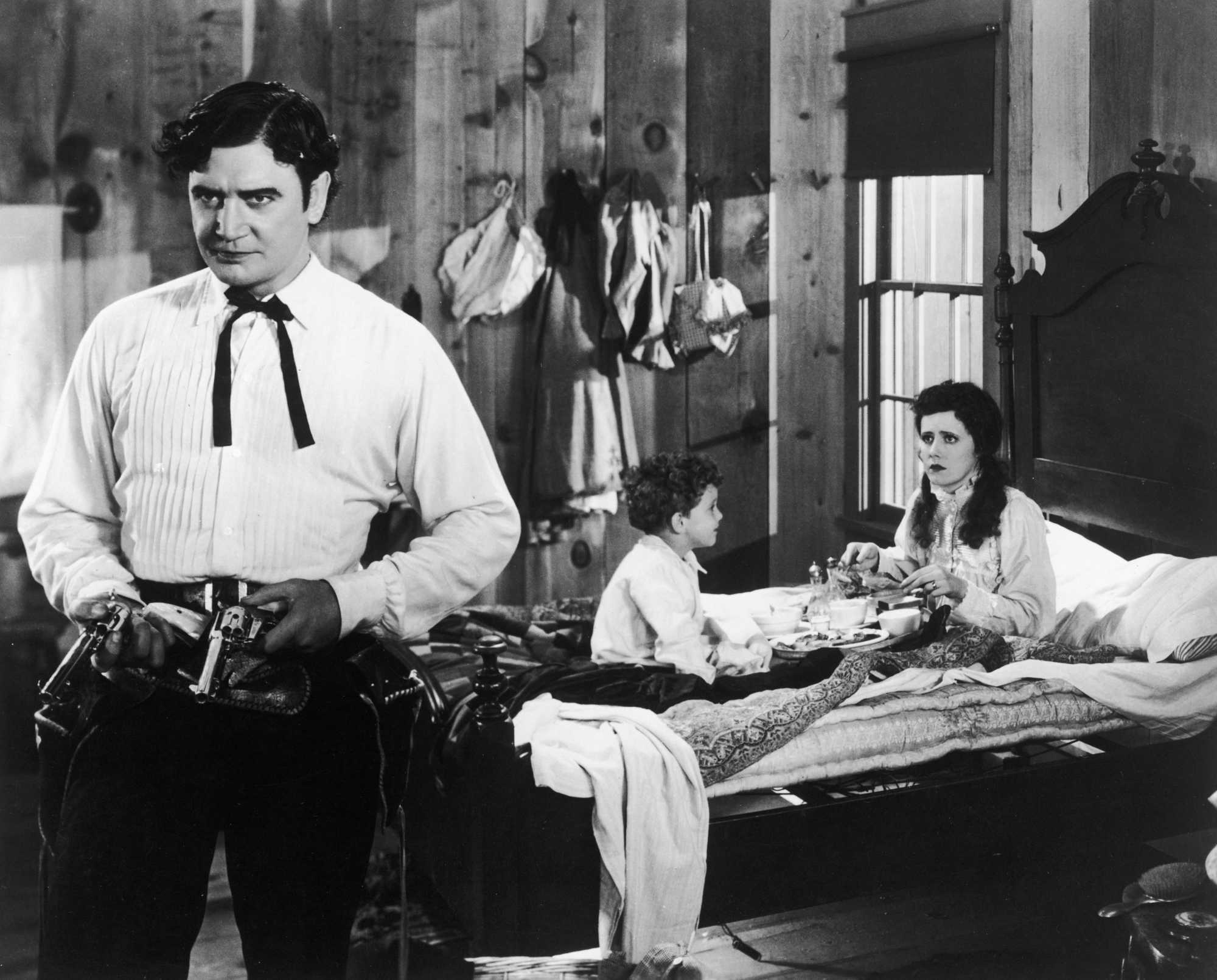 'Cimarron' Richard Dix as Yancey Cravat, Irene Dunne as Sabra Cravat, and Douglas Scott as Toddler Cim. Dix is standing with two pistols, while Dunne and Scott sit on the bed behind him.