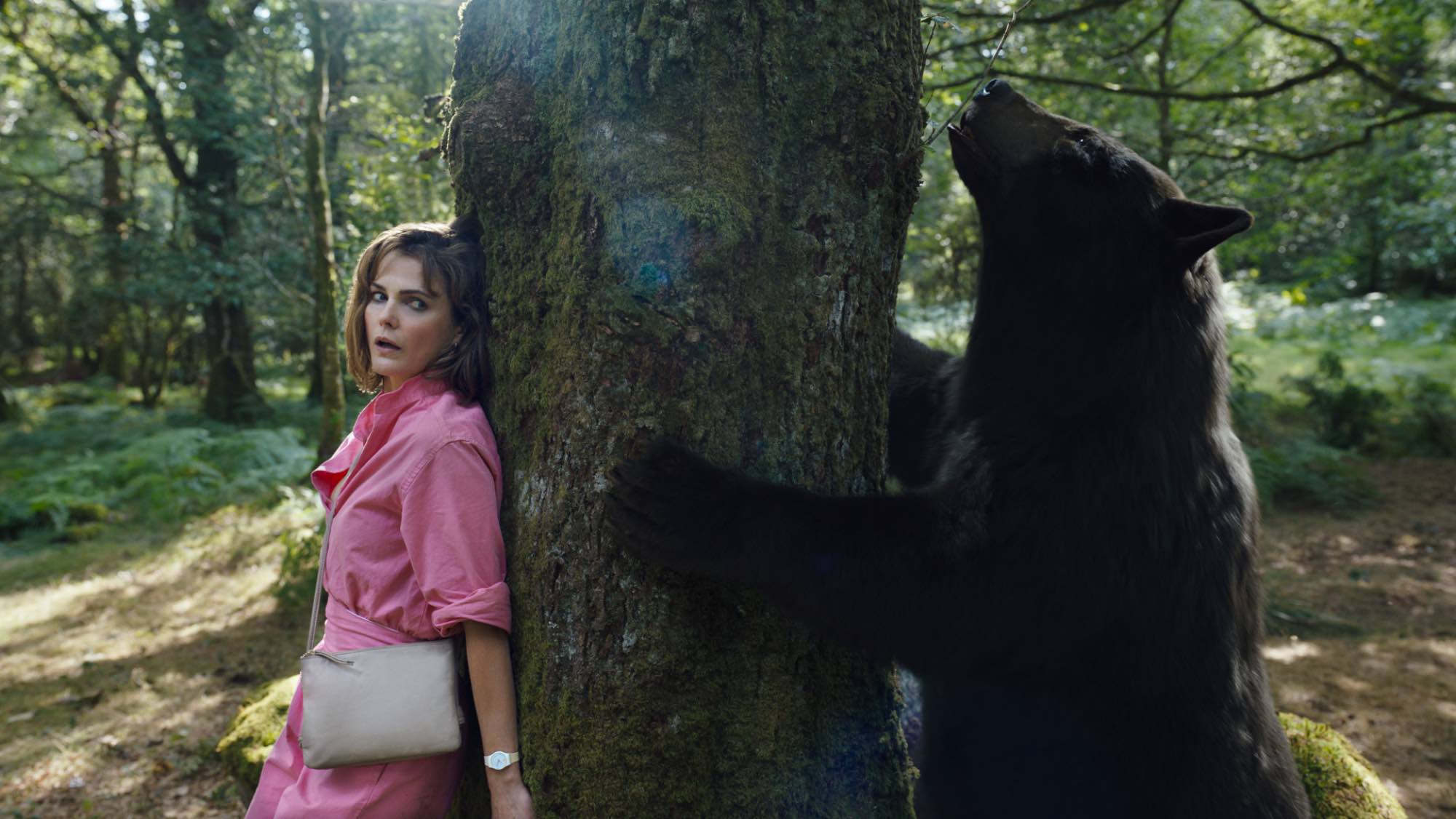 'Cocaine Bear' Keri Russell as Sari wearing pink while hiding behind a tree from a black bear on the other side