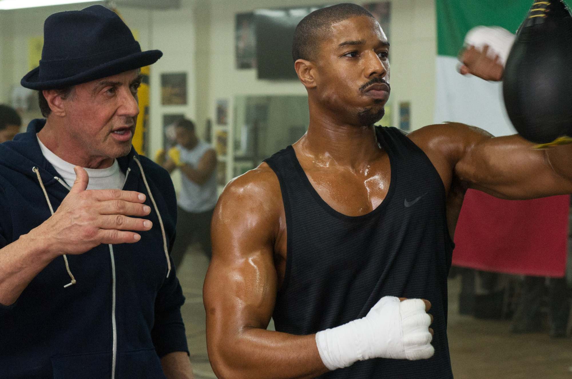 'Creed' Sylvester Stallone as Rocky and Michael B. Jordan as Adonis Creed. Rocky is standing behind Adonis in a boxing gym, punching a hanging bag.