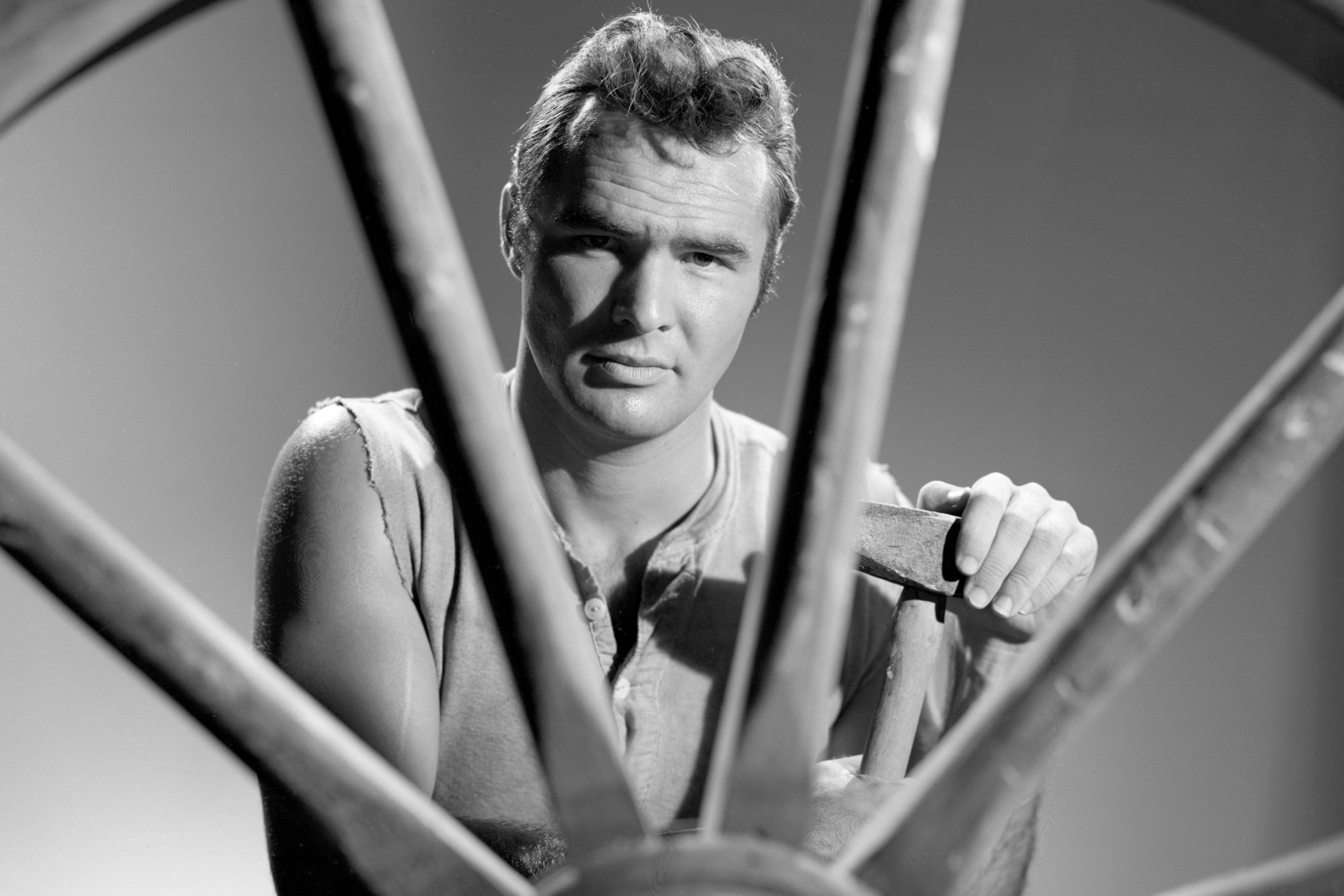 'Gunsmoke' actor Burt Reynolds as Quint Asper holding a blacksmith tool sitting behind a wheel