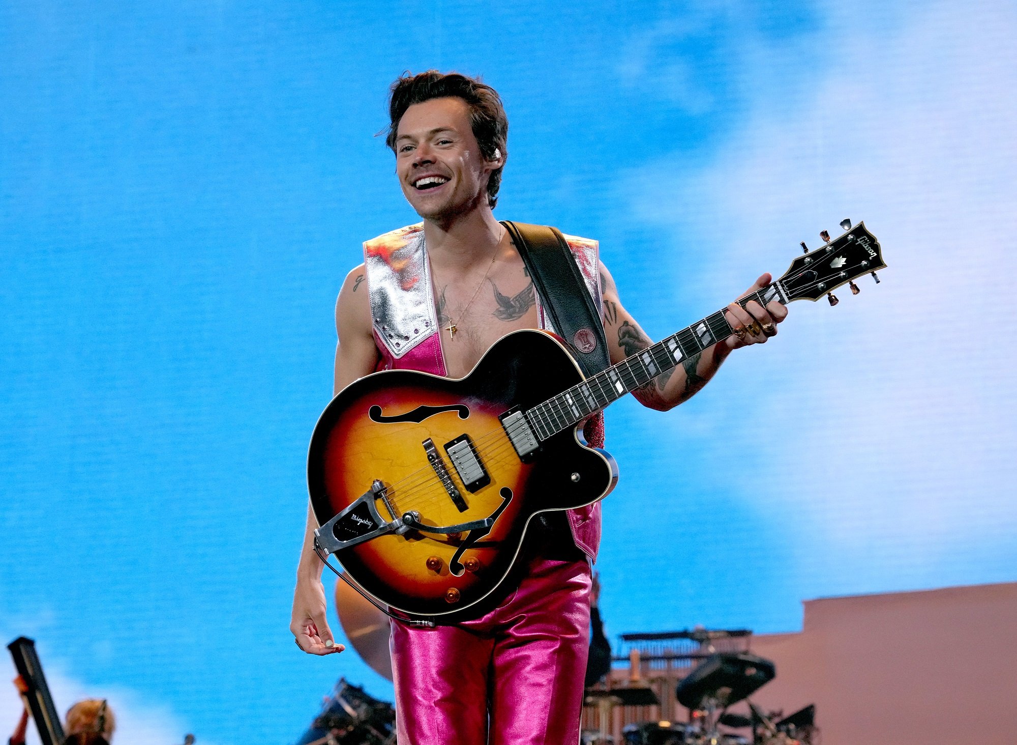 Harry Styles smiles on stage while holding a guitar