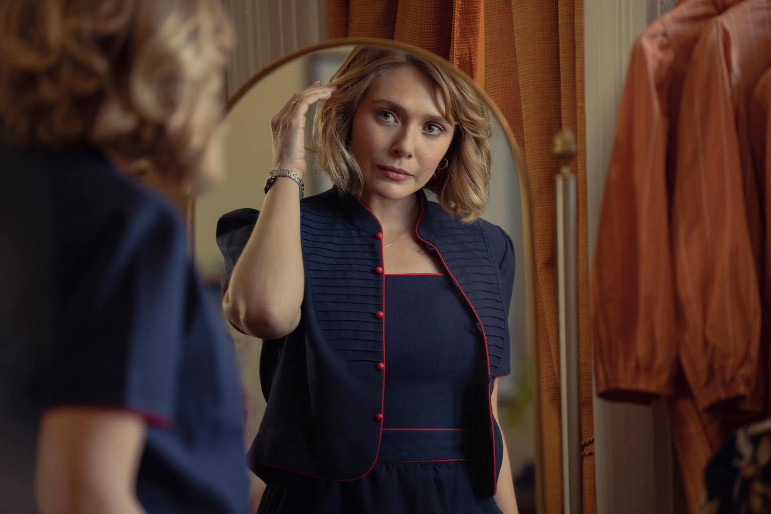 HBO Max 'Love and Death' stars Elizabeth Olsen as Candy Montgomery, seen here wearing a navy sweater and dress while looking into a mirror in a production still.