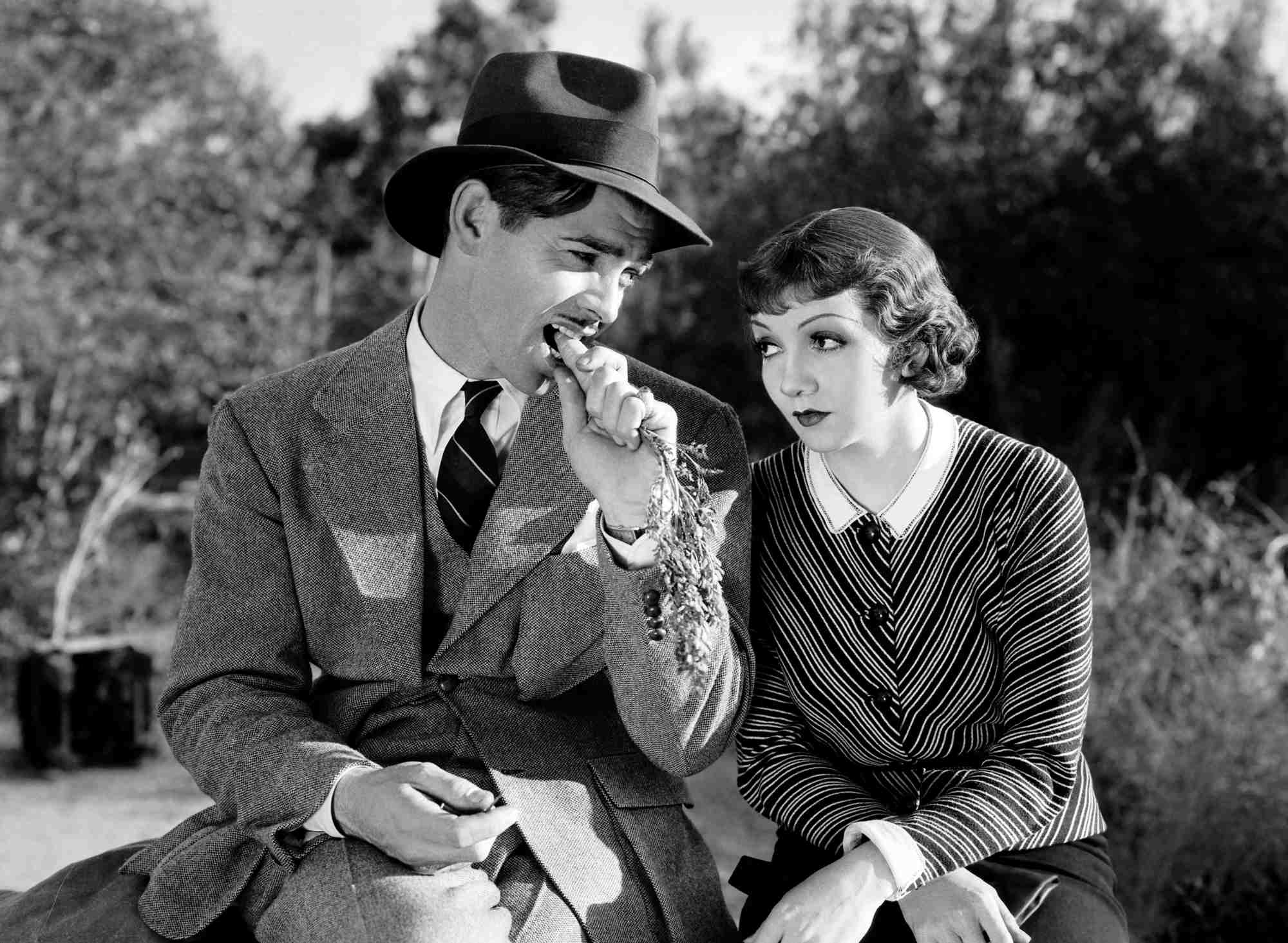 'It Happened One Night' Clark Gable as Peter Warne and Claudette Colbert as Ellie Andrews sitting next to each other in nature, with Gable chewing on a carrot