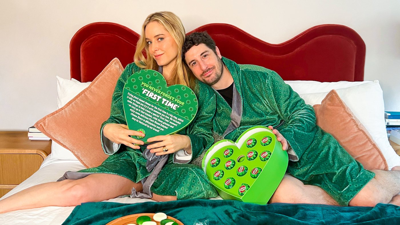 Jenny Mollen and Jason Biggs sit on a bed wearing green robes and a green heart shaped box with cheese