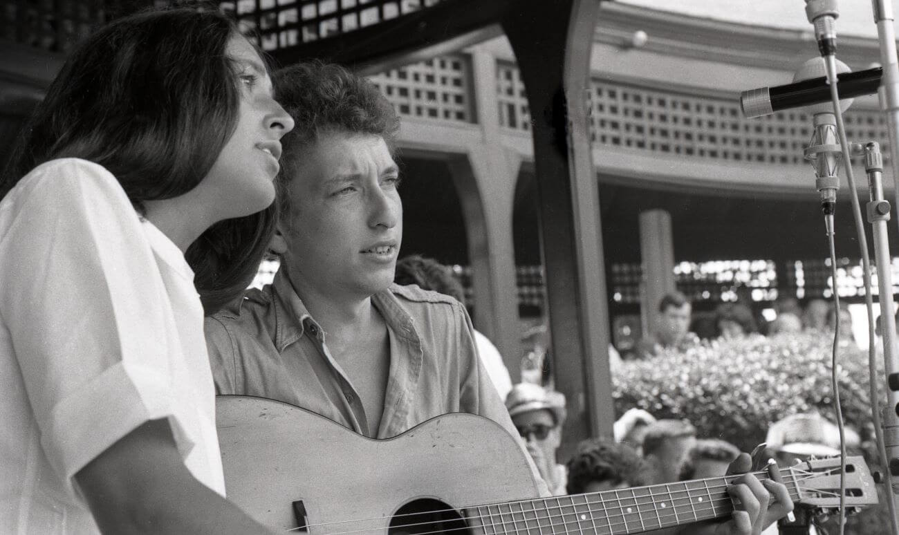 A black and white picture of Bob Dylan holding a guitar and sitting onstage with Joan Baez.