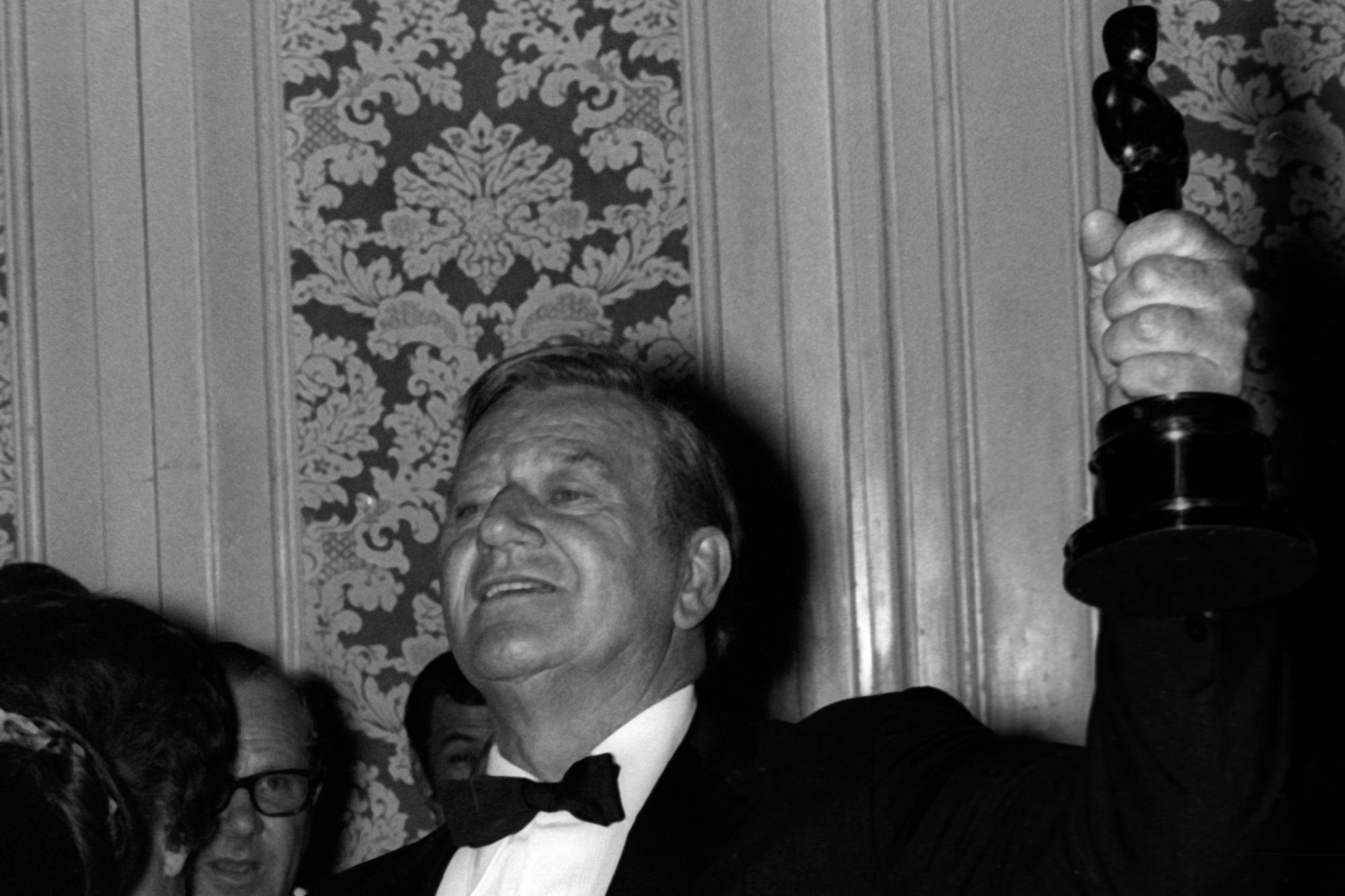 John Ford collaborator John Wayne holding an Oscar in the air, photographed from below