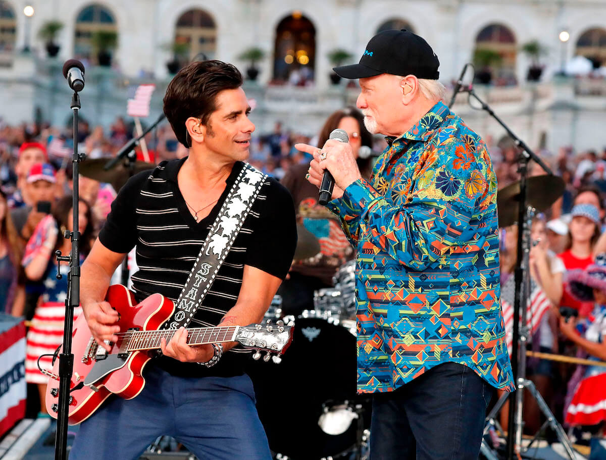 John Stamos sitting with a guitar looking at Mike Love singing into a microphone