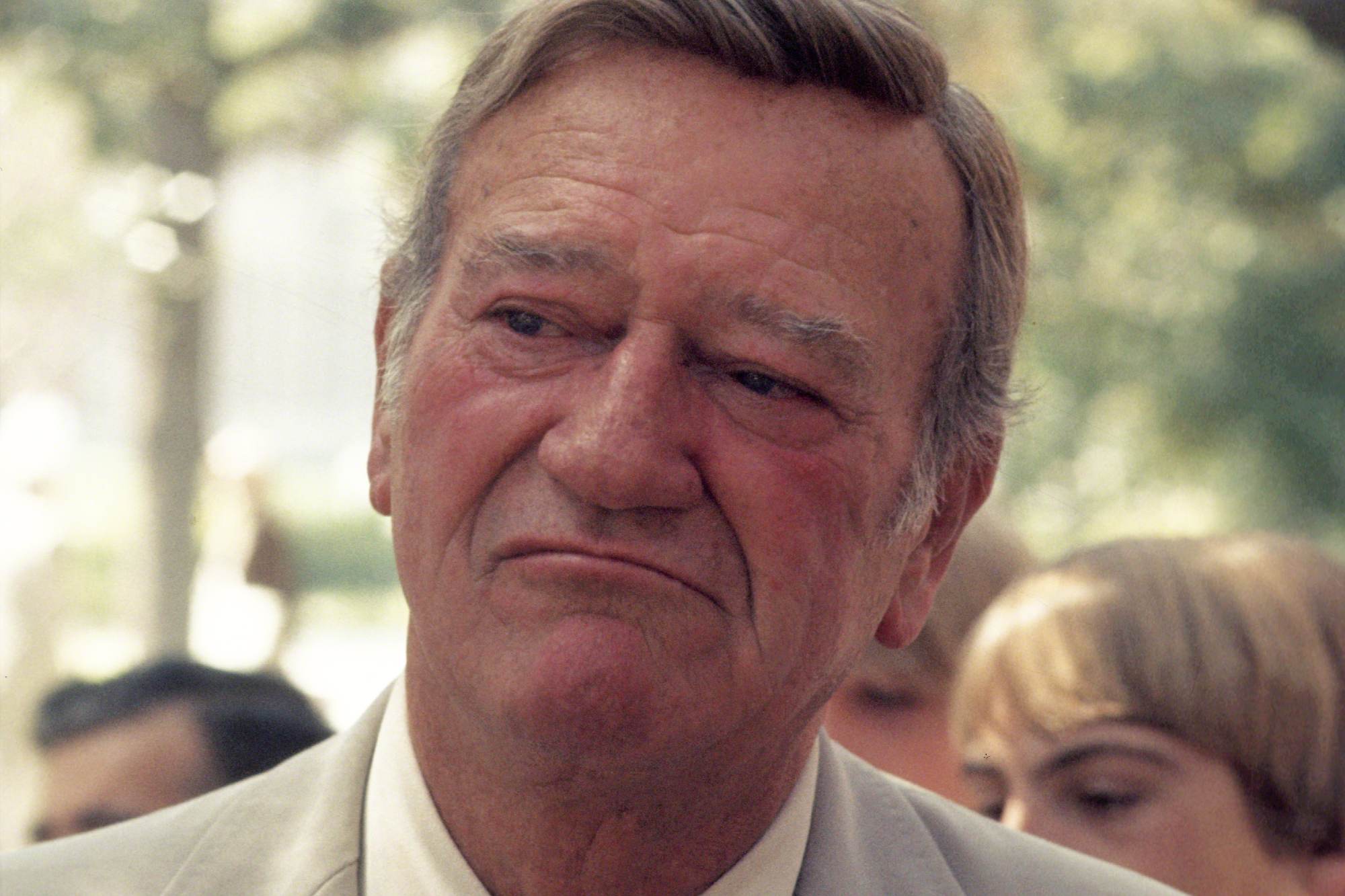 John Wayne, an actor from America. He's wearing a suit, looking to the side, with a frown.