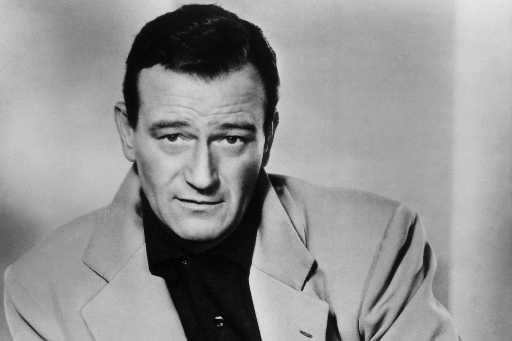 John Wayne, who starred in Western and war movies. Black-and-white picture of Wayne looking at the camera, while wearing a suit jacket.