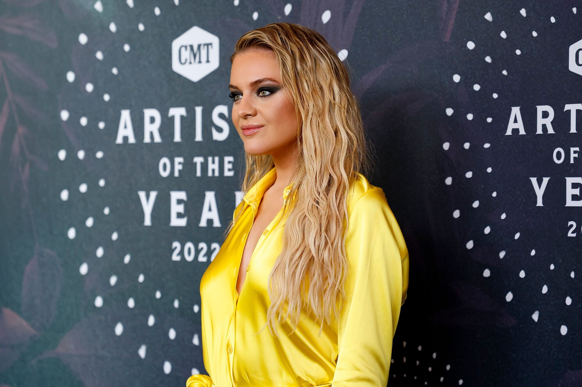 Kelsea Ballerini wears a long-sleeved yellow outfit in front of a black backdrop that reads 'CMT Artist of the Year 2022'