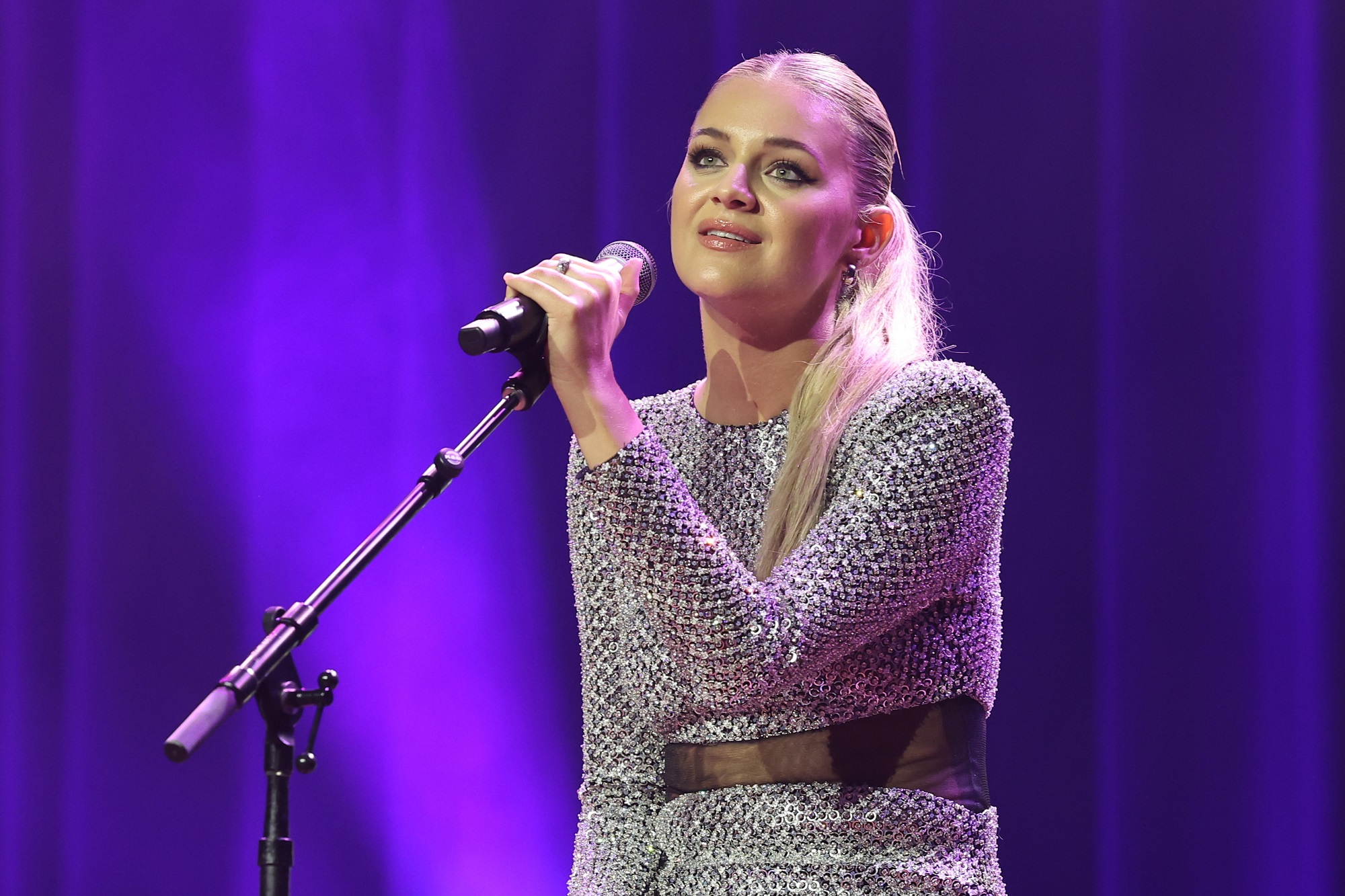 Kelsea Ballerini holds a microphone onstage while wearing a silver dress