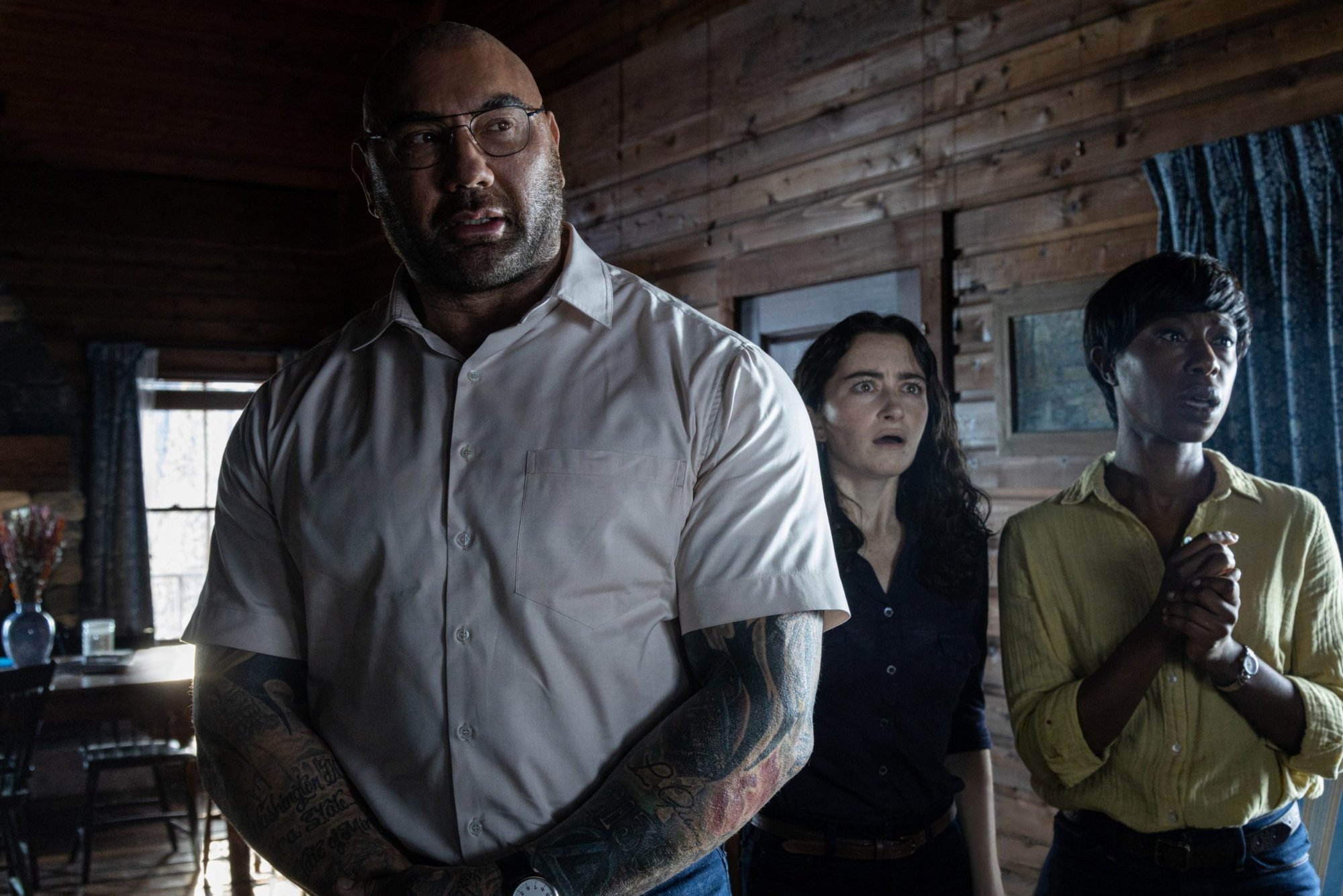 'Knock at the Cabin' Dave Bautista as Leonard, Abby Quinn as Ardiane, and Nikki Amuka-Bird as Sabrina standing in wood cabin looking shocked