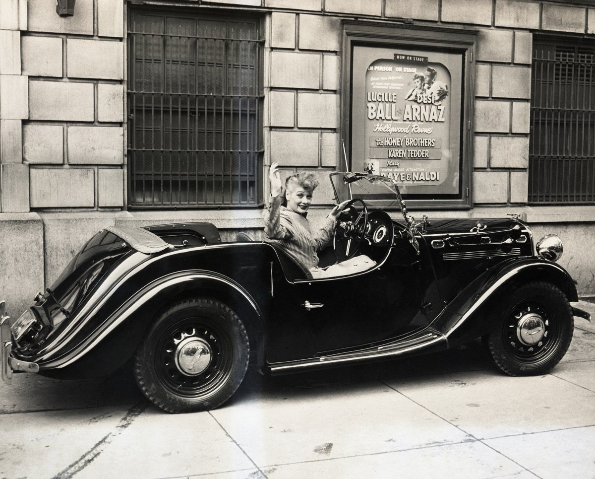 Lucille Ball is seen in a sports roadster in front of a poster advertising one of her projects