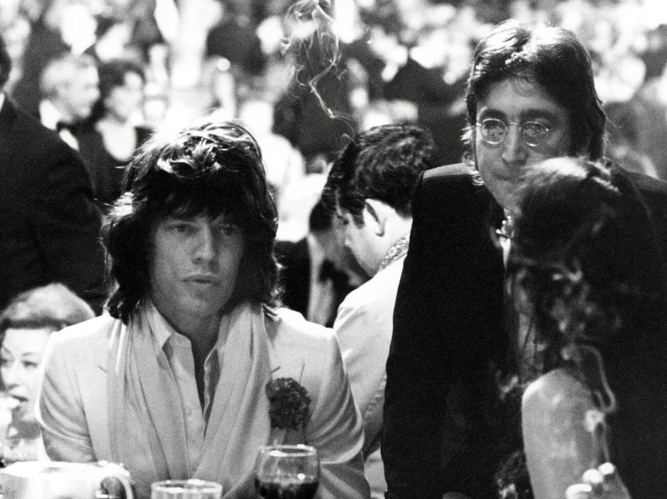 A black and white picture of Mick Jagger and John Lennon sitting next to each other in a crowded room.