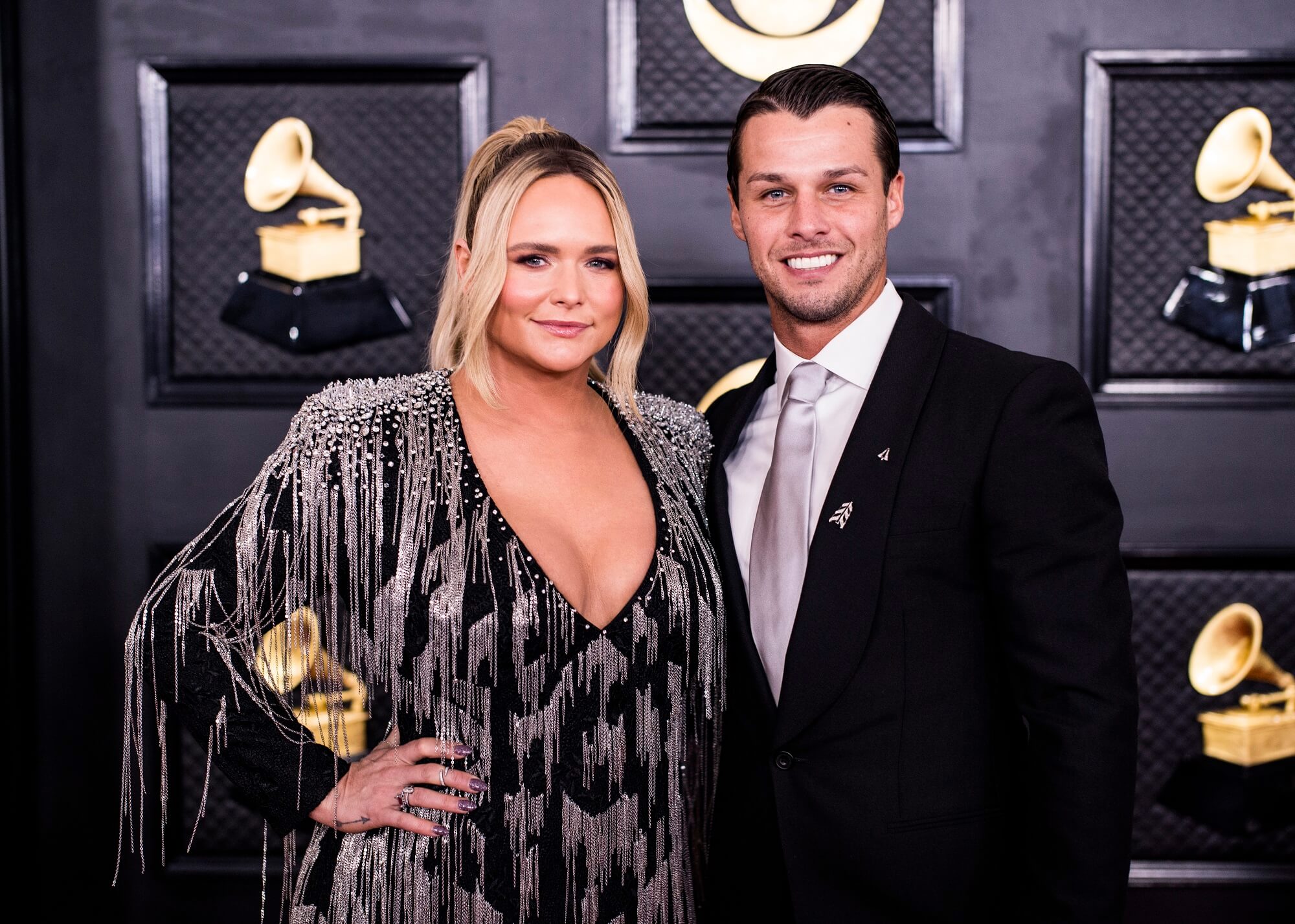 Miranda Lambert and Brendan McLoughlin smile together in front of a black backdrop with gold Grammy trophies on it
